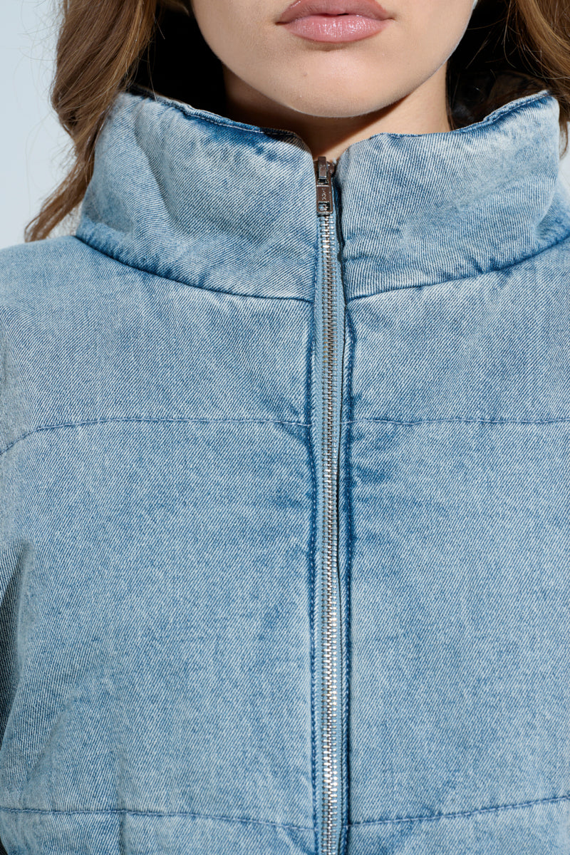 Close-up of someone wearing the CHARLI CROPPED DENIM PUFFER, focusing on its textured design, high collar, and silver zipper. The person's face is partially visible.