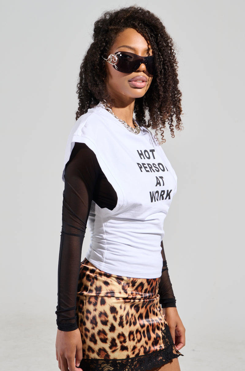 A person wearing sunglasses, a "HOT PERSON AT WORK LONG SLEEVE GRAPHIC SHIRT" layered under a black graphic long-sleeve shirt and paired with a leopard print skirt. With curly hair, they look confidently towards the camera.