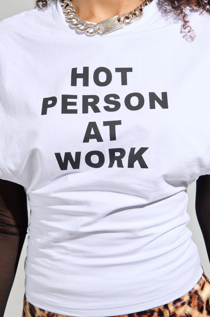 A person dressed in a white "HOT PERSON AT WORK" long sleeve graphic shirt, featuring bold black text, paired with a chunky silver chain necklace and patterned pants. They are standing against a plain background, exuding confidence.