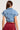 A person with long brown hair is shown from the back. They are wearing the FOREVER THAT GIRL DENIM BUTTON UP SHIRT, a short-sleeved, blue wash denim jacket with a collar, styled like a crop top, and a high-waisted, pleated burgundy skirt. The background is plain and neutral.