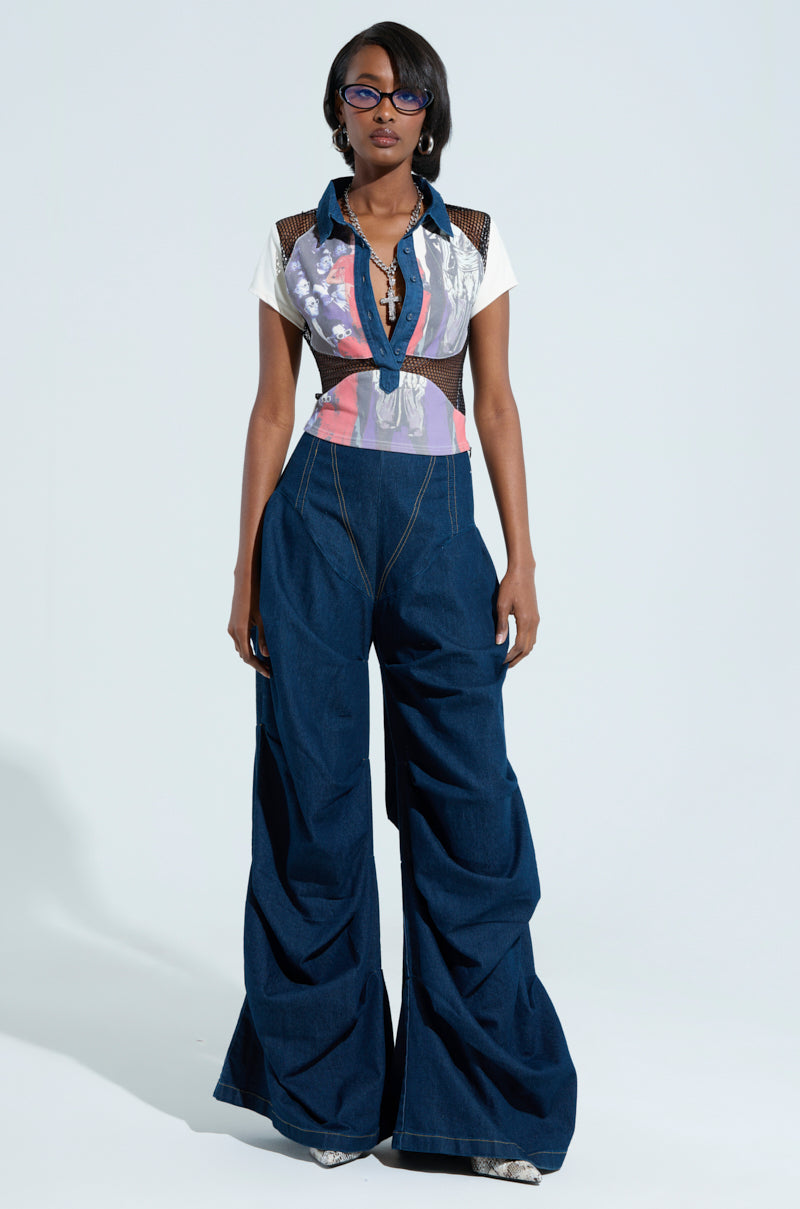 A person wearing the Edgy Diva Short Sleeve Netted Tshirt and dark wide-leg jeans stands confidently. They are accessorized with glasses and a necklace, posing against a plain light background.
