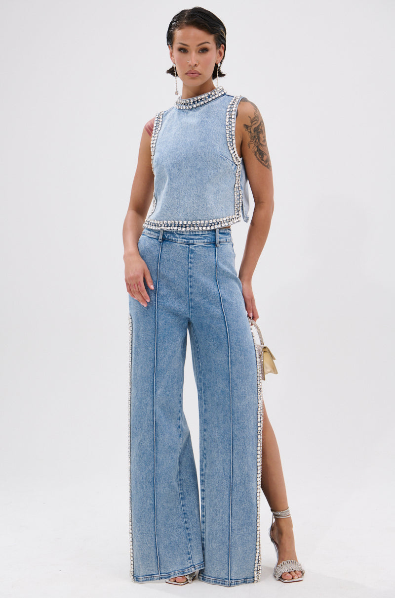 A woman with braided hair and tattoos poses in POPPING OUT RHINESTONE DENIM PANTS—wide-leg, high-rise jeans adorned with sparkling embellishments—paired with a sleeveless light blue top and open-toe heels against a plain white background.