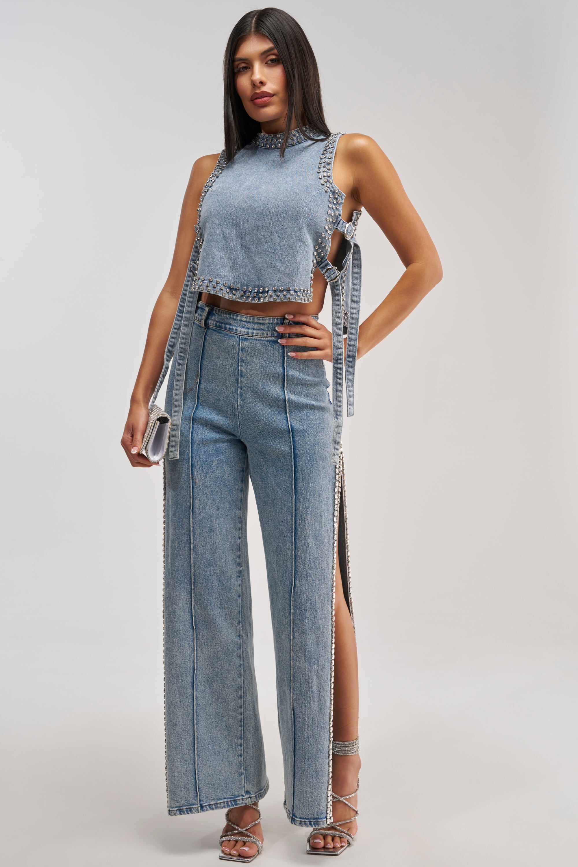 A woman stands against a plain light background wearing a sleeveless, light denim crop top with side ties paired with the POPPING OUT RHINESTONE DENIM PANT, accessorized with a silver handbag and strappy silver heels.