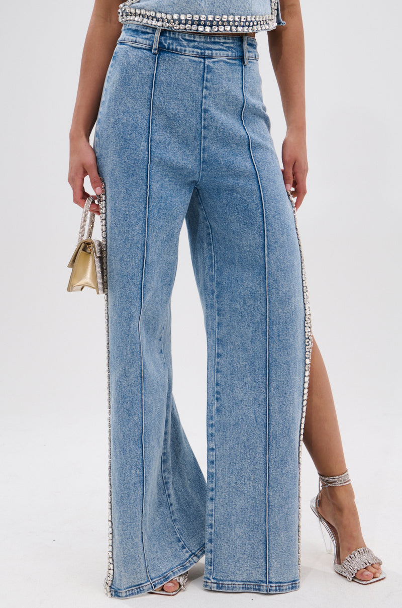 Someone wears POPPING OUT RHINESTONE DENIM PANT—high-rise, wide-leg blue jeans with rhinestone embellishments on the seams—paired with silver heeled sandals and a small gold handbag. The top half of their body isn't visible.