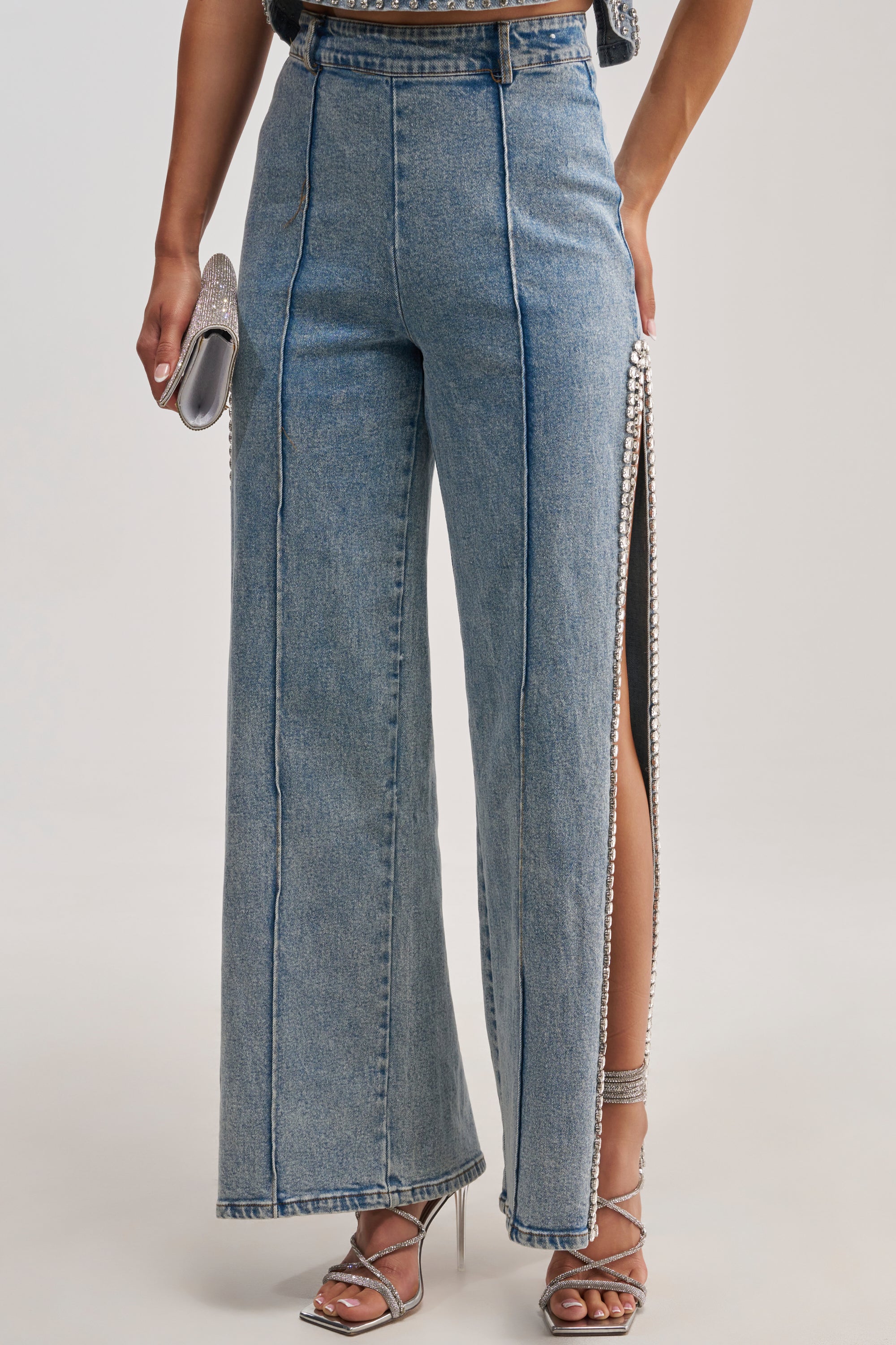 Wearing the POPPING OUT RHINESTONE DENIM PANT—high-waisted, straight-leg jeans in a blue wash featuring front seams and bold high side slits embellished with rhinestones—the person holds a silver clutch and wears strappy heels. Only their lower body is visible.