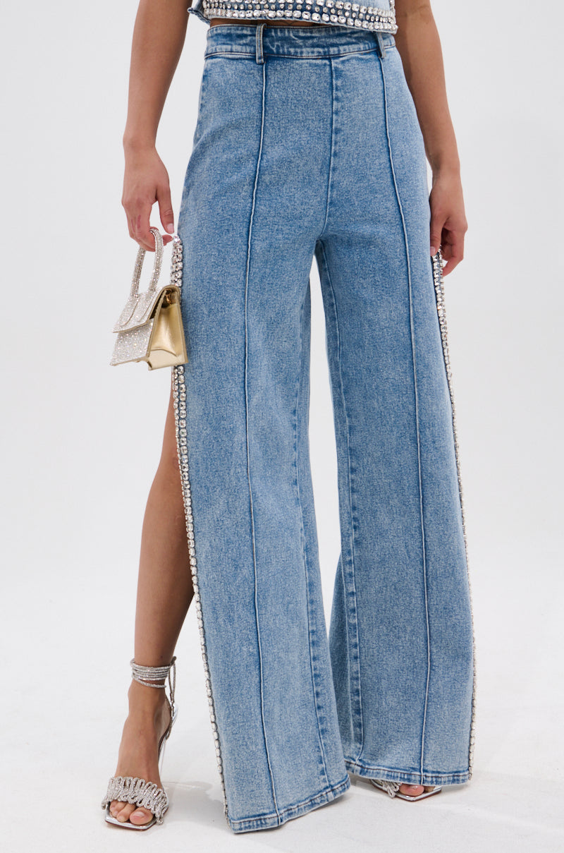 A person wears the POPPING OUT RHINESTONE DENIM PANT featuring a high-rise waist and sparkling seam embellishments, styled with a small glittery handbag and silver jeweled-strap high-heeled sandals.