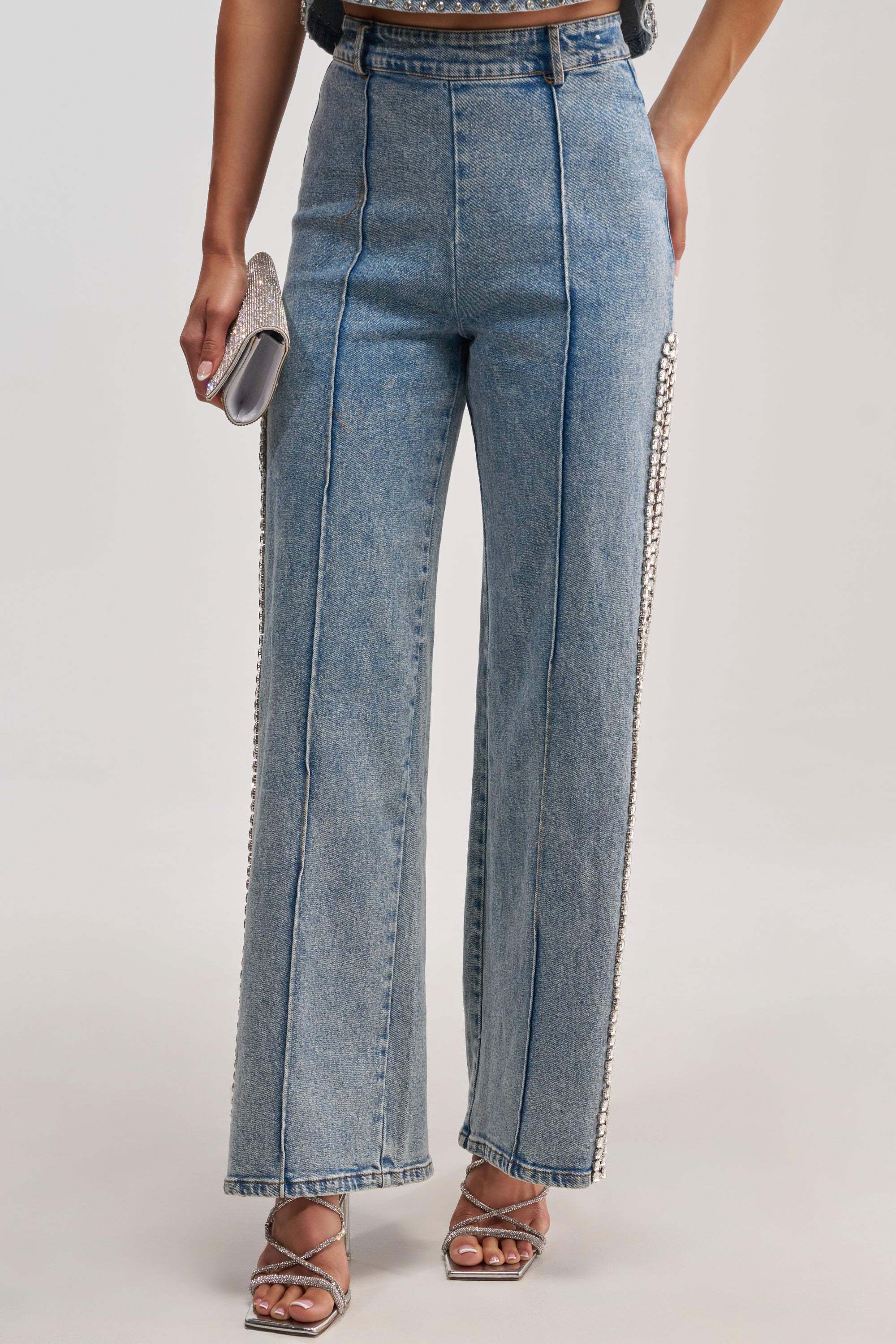 A person wearing the POPPING OUT RHINESTONE DENIM PANT, a pair of high-waisted, wide-leg blue jeans with sparkling rhinestone embellishments along the side seams, paired with silver heeled sandals and holding a silver clutch.