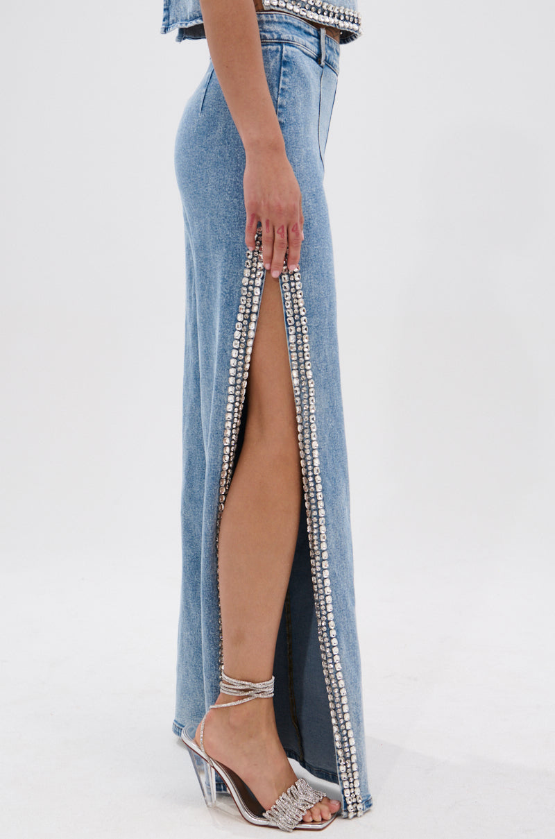 A person wears the POPPING OUT RHINESTONE DENIM PANT featuring blue denim, rhinestone embellishments, high side slits, and silver strappy heels. The background is plain white.