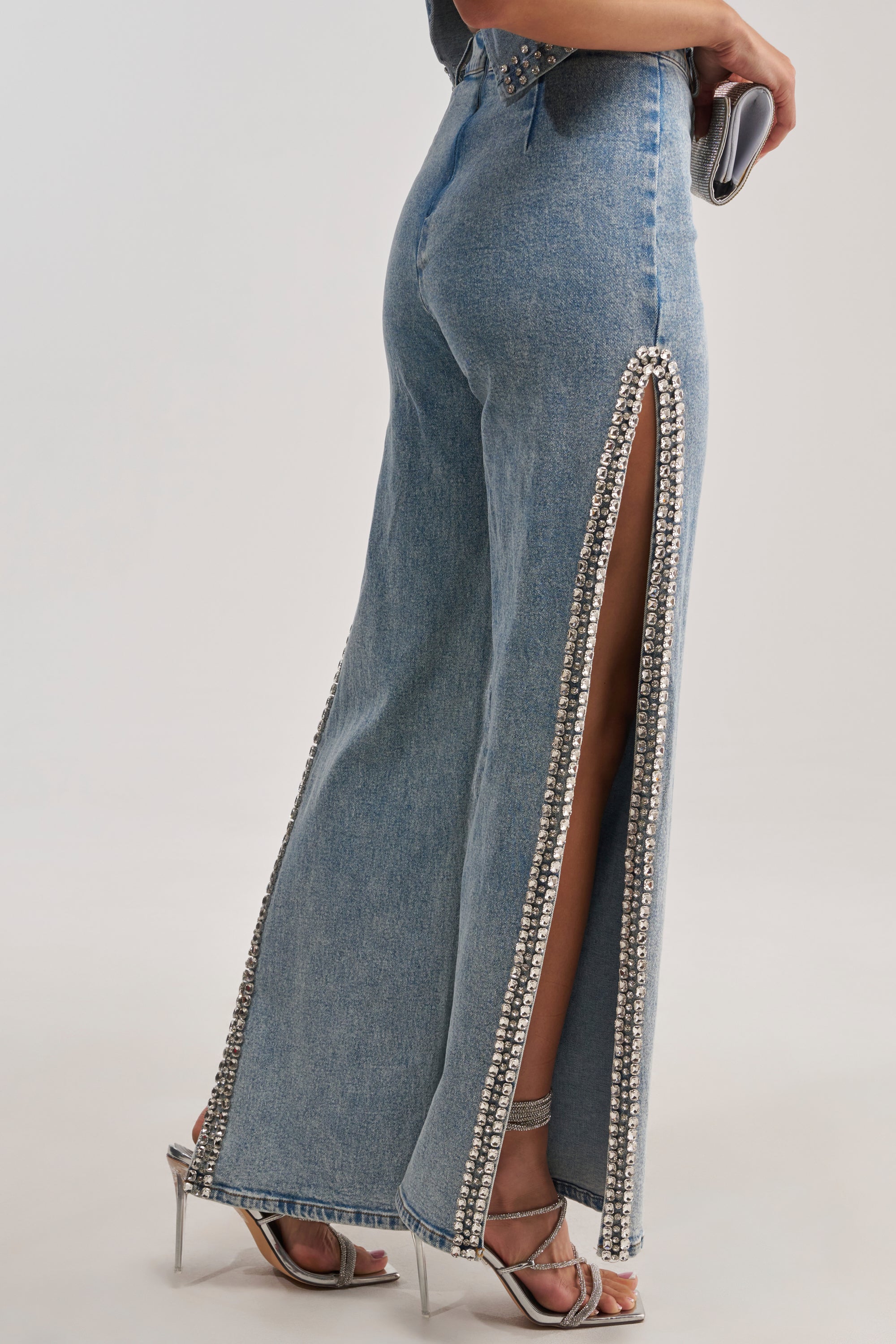 Person wearing the POPPING OUT RHINESTONE DENIM PANT—a wide-leg blue denim style with high side slits embellished with rhinestones—paired with a small clutch and metallic high-heeled sandals. Only the lower body is visible.