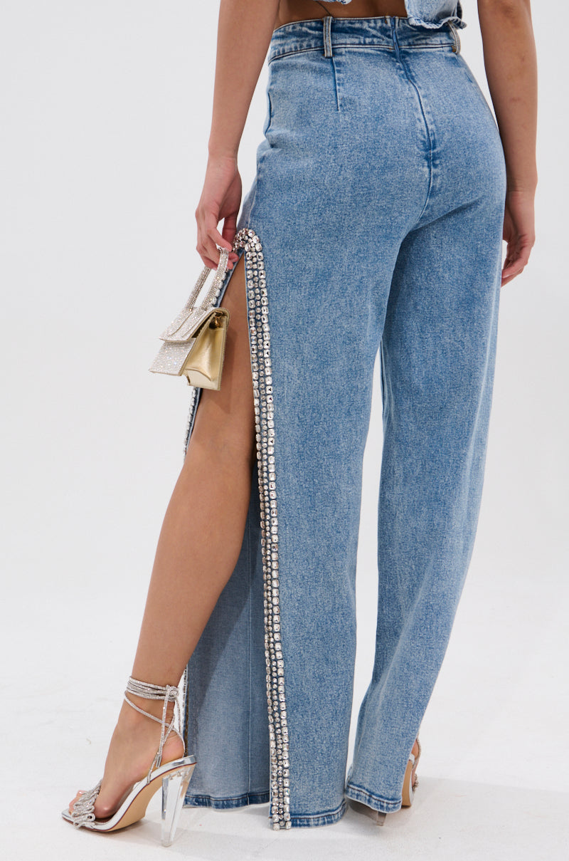 Wearing the POPPING OUT RHINESTONE DENIM PANT with high-rise fit and side slits, paired with strappy heels and a small gold and silver purse, the photo showcases the sparkling rhinestone embellishments from behind.