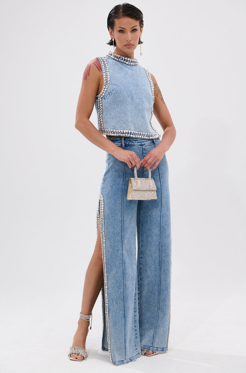 A woman poses in a sleeveless, high-neck denim top and the POPPING OUT RHINESTONE DENIM PANT, featuring high-rise waist, side slits, and sparkling embellishments. She accessorizes with a silver handbag and strappy heels with crystal details.