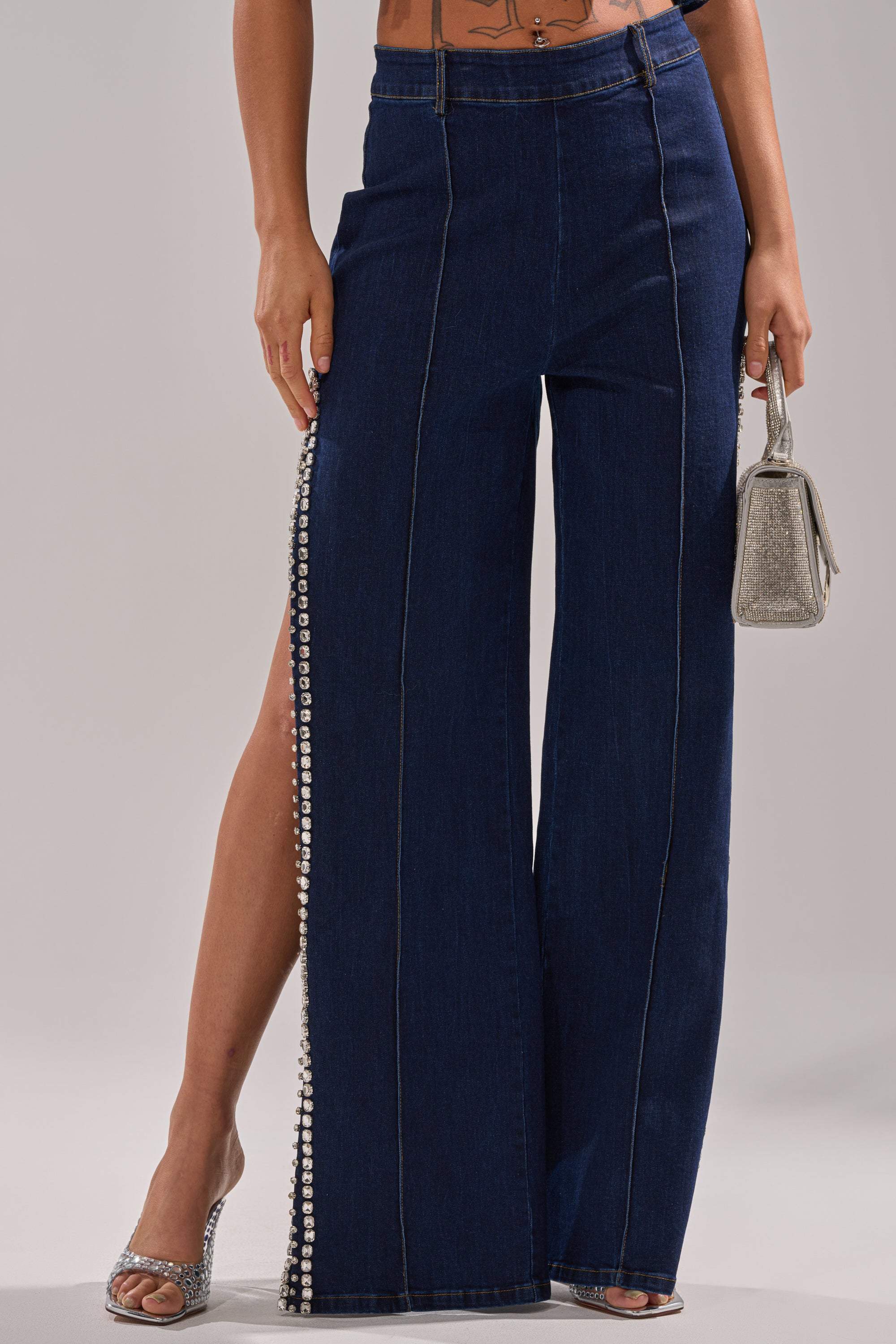 A person wearing the POPPIN OUT RHINESTONE DENIM PANT IN DARK WASH DENIM with rhinestone-trimmed side slits, paired with silver open-toe heels and carrying a small glittery silver handbag.