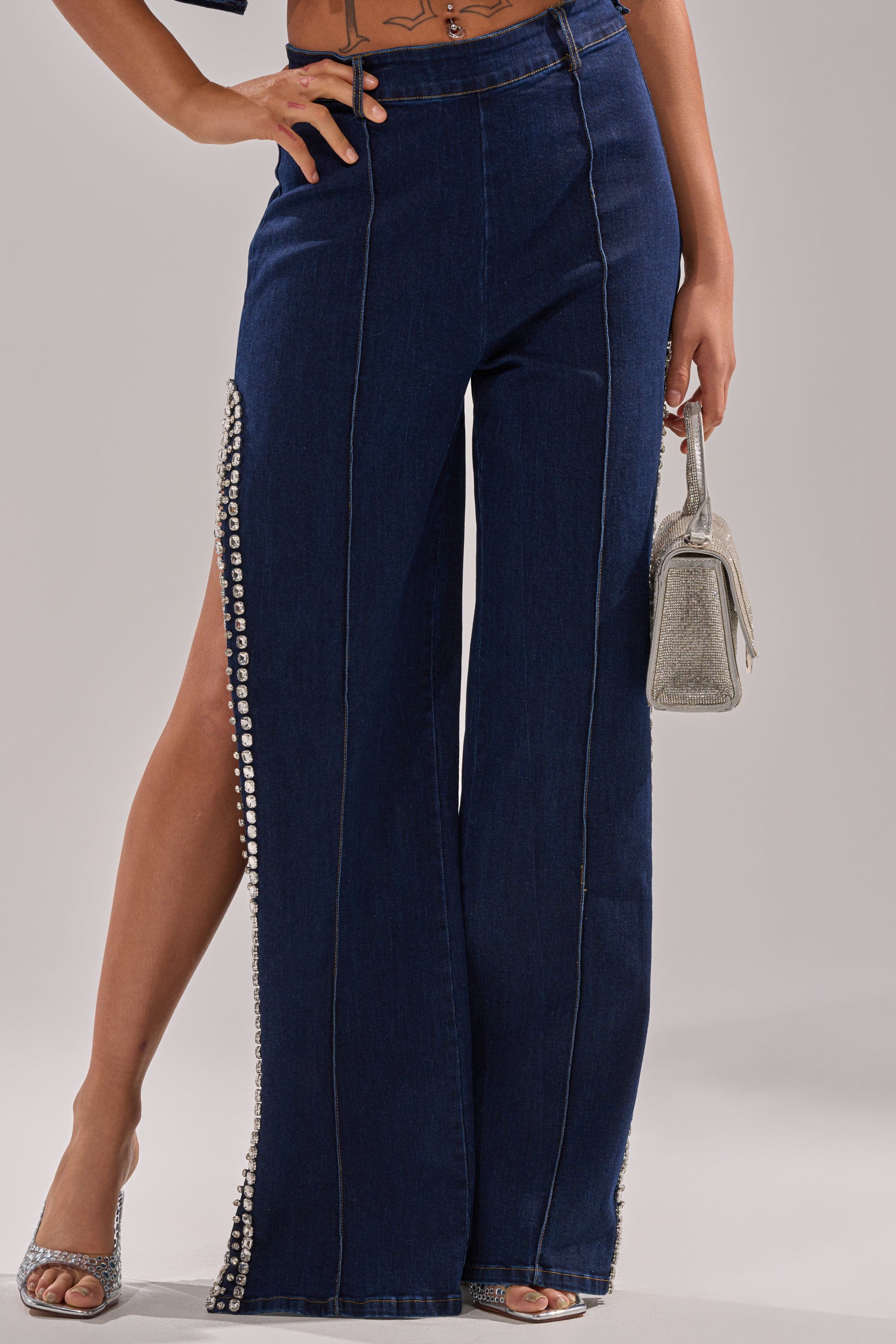 A woman wears the POPPIN OUT RHINESTONE DENIM PANT in dark wash, featuring high-rise flare legs and rhinestone-adorned side slits. She carries a sparkly silver handbag, dons matching heels, with her midriff and one arm partly visible.