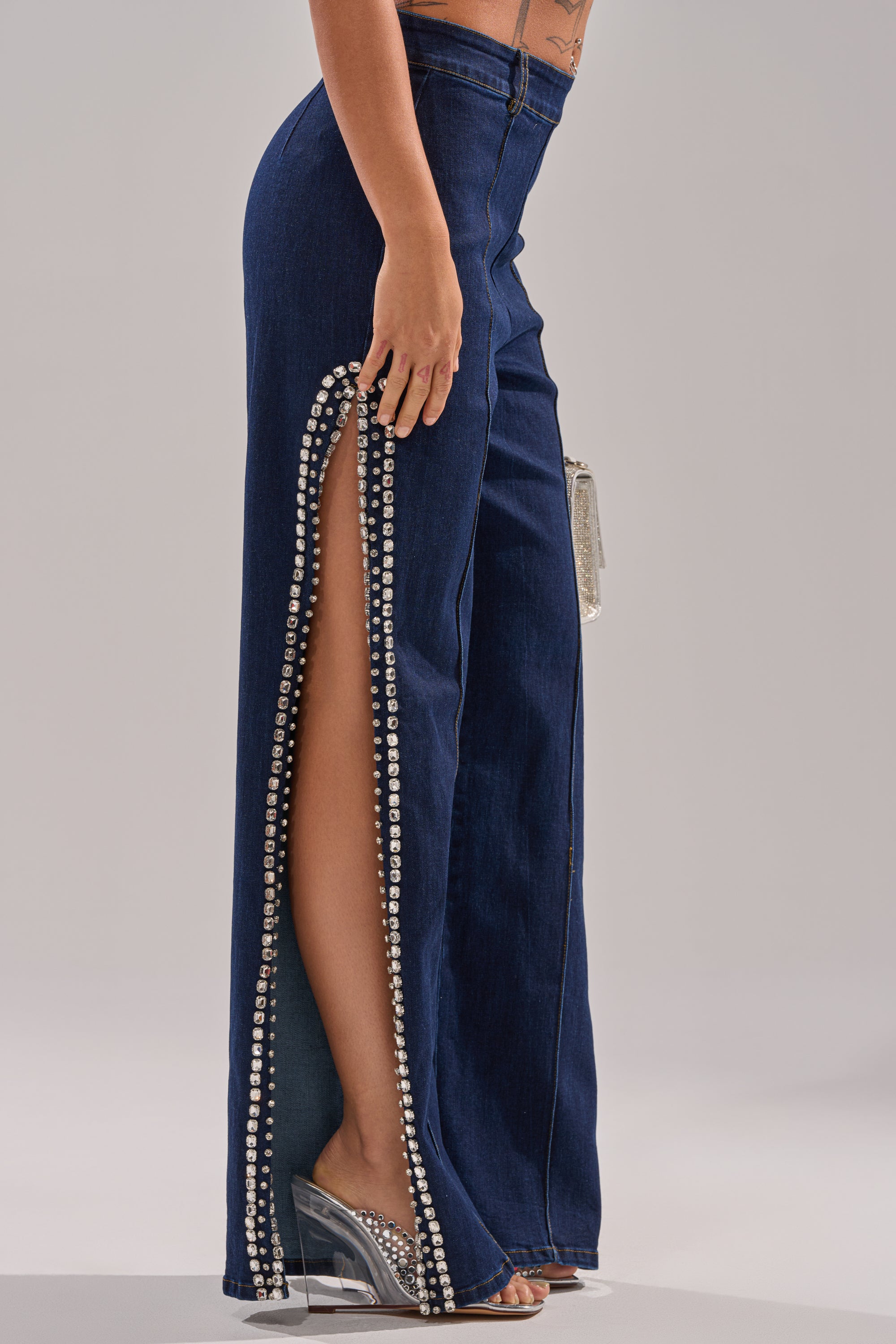 A person models the POPPIN OUT RHINESTONE DENIM PANT IN DARK WASH DENIM, featuring a high-rise flare style with a long rhinestone-adorned side slit, paired with clear high heels against a light gray background.