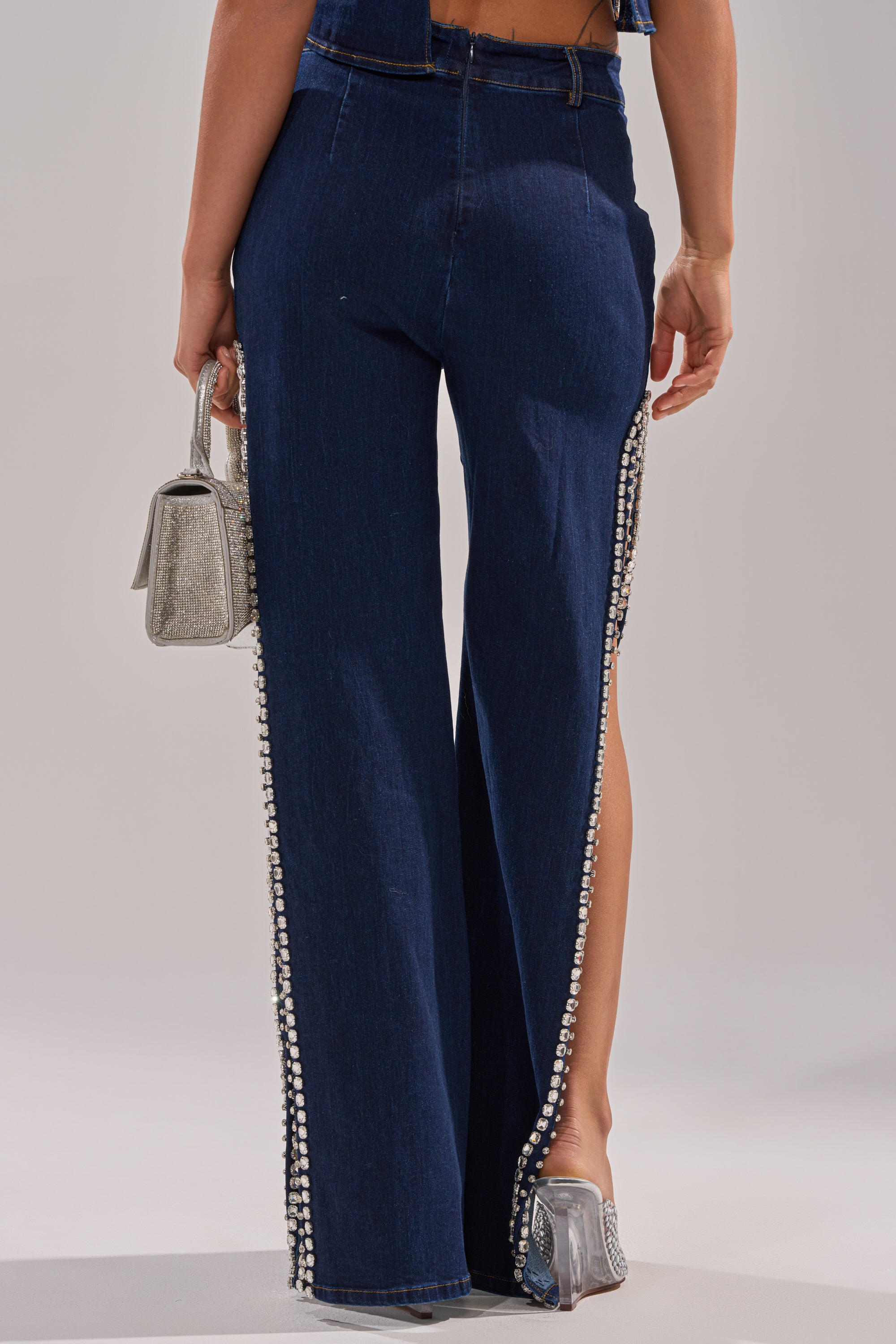 A person wears the POPPIN OUT RHINESTONE DENIM PANT IN DARK WASH DENIM, featuring pearl side embellishments, paired with a silver mini handbag and silver high heels. The photo captures them from behind.