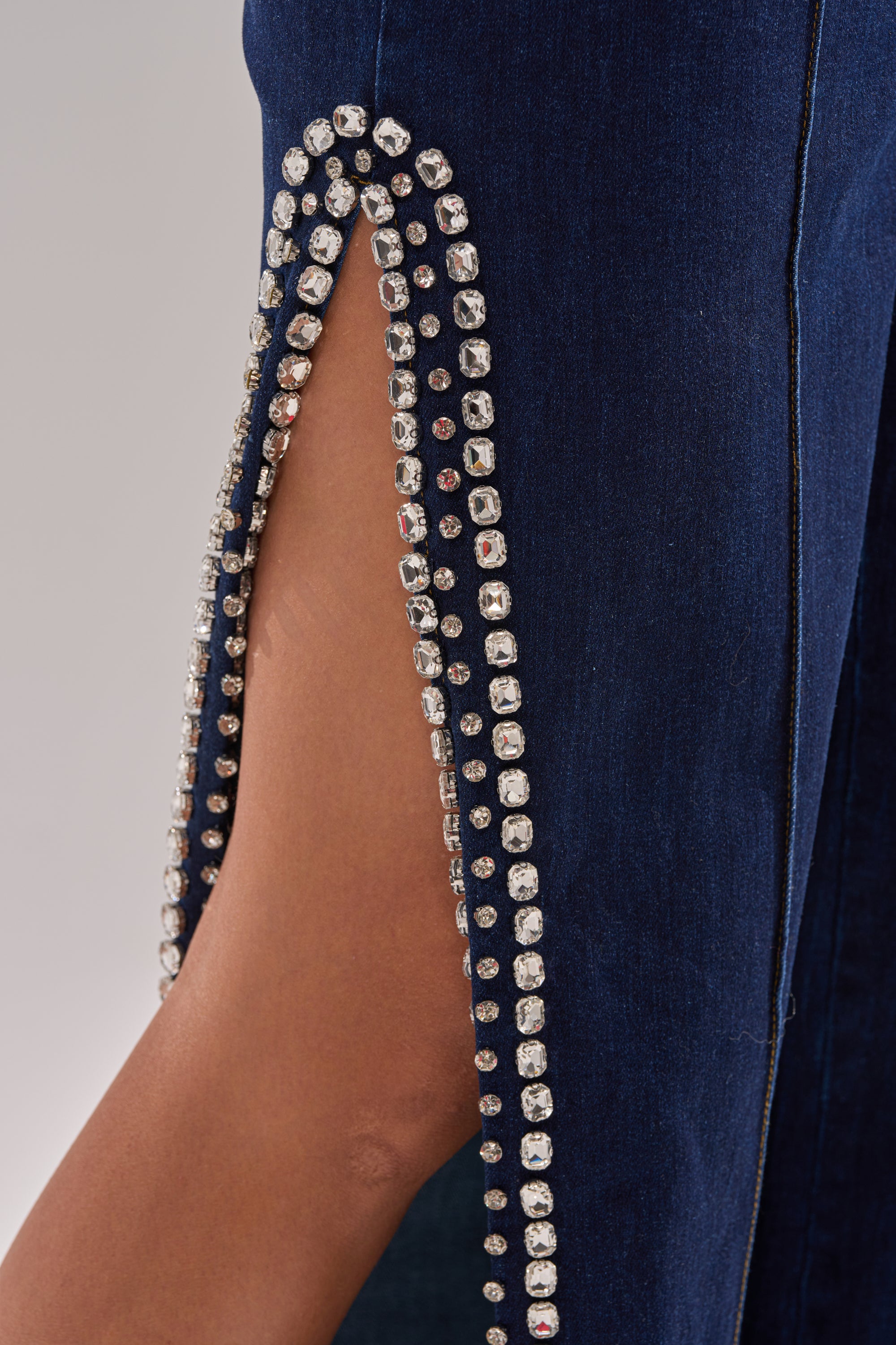 Close-up of a person wearing the POPPIN OUT RHINESTONE DENIM PANT IN DARK WASH DENIM, featuring a high-rise flare fit with a thigh-high slit edged in double rows of sparkling rhinestones, revealing some skin underneath.