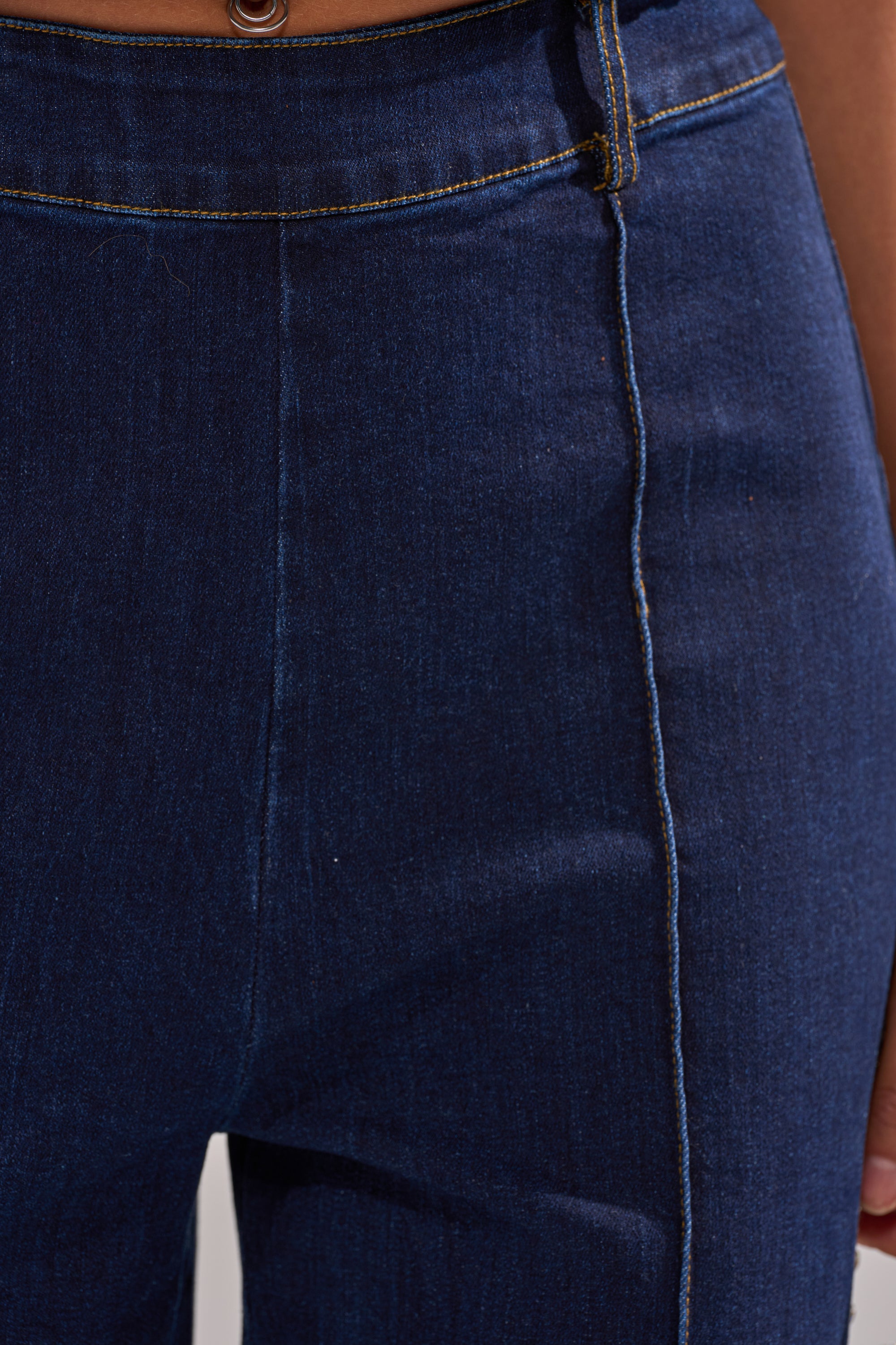 Close-up of a person wearing the POPPIN OUT RHINESTONE DENIM PANT IN DARK WASH DENIM, featuring high-rise flare style, yellow contrast stitching, and a visible waistband button.