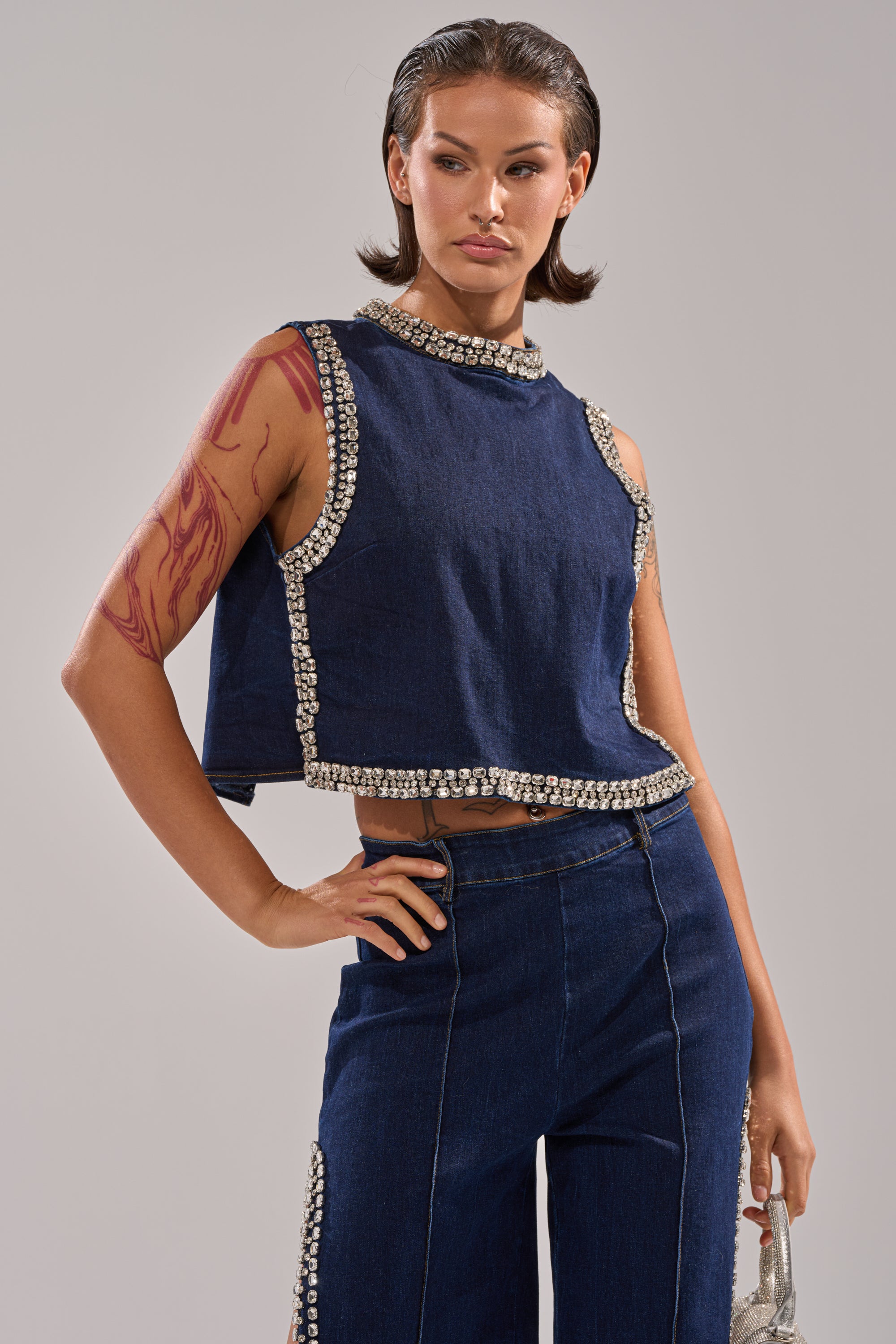 A woman wearing the POPPIN OUT RHINESTONE DENIM BLOUSE IN DARK WASH DENIM and matching pearl-embellished wide-leg pants poses with one hand on her hip, showcasing a red abstract tattoo on her upper arm. 0