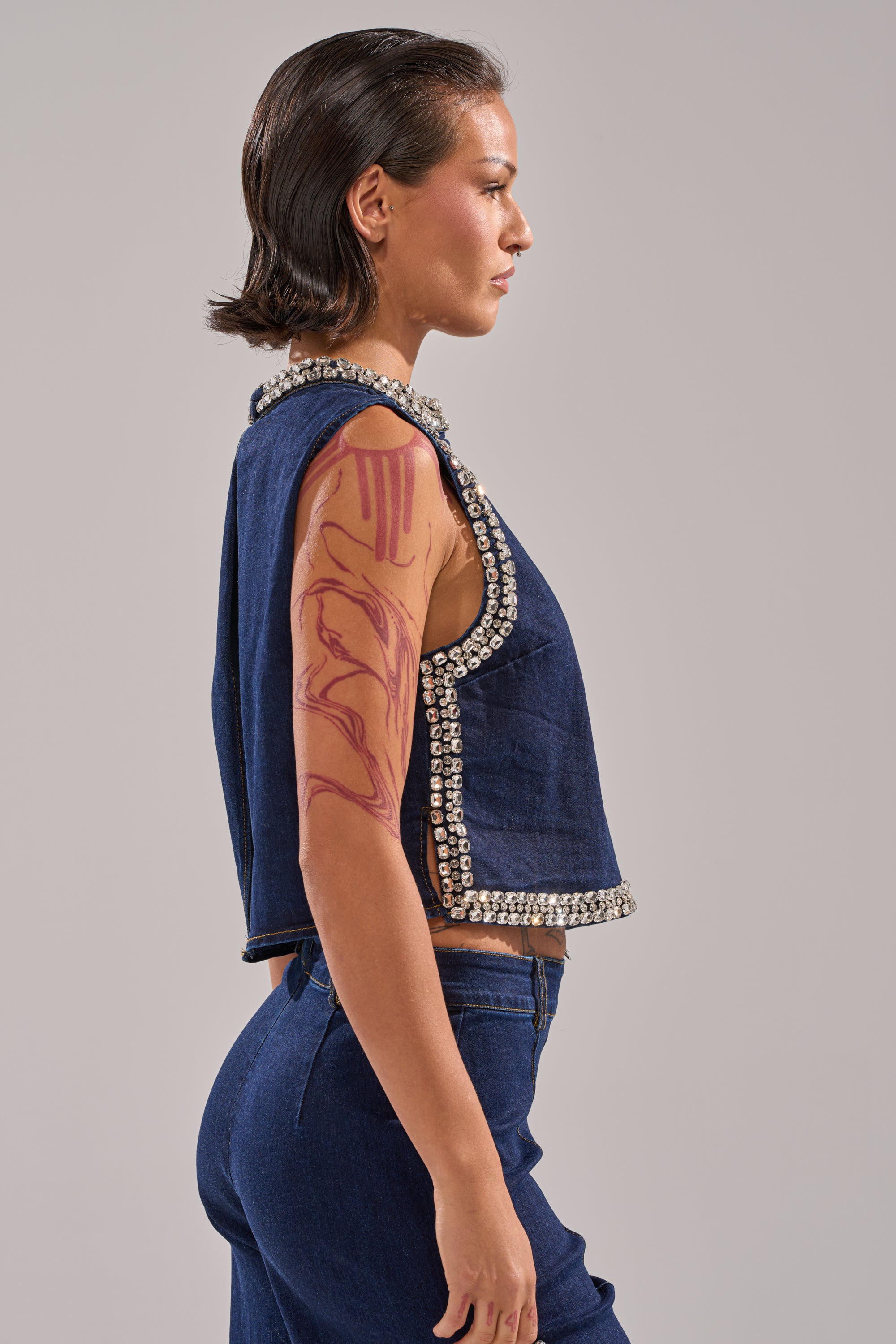 A woman with short, dark hair stands in profile, wearing the POPPIN OUT RHINESTONE DENIM BLOUSE IN DARK WASH DENIM and matching jeans. She has bold red upper arm body art. The background is plain and light-colored. 3