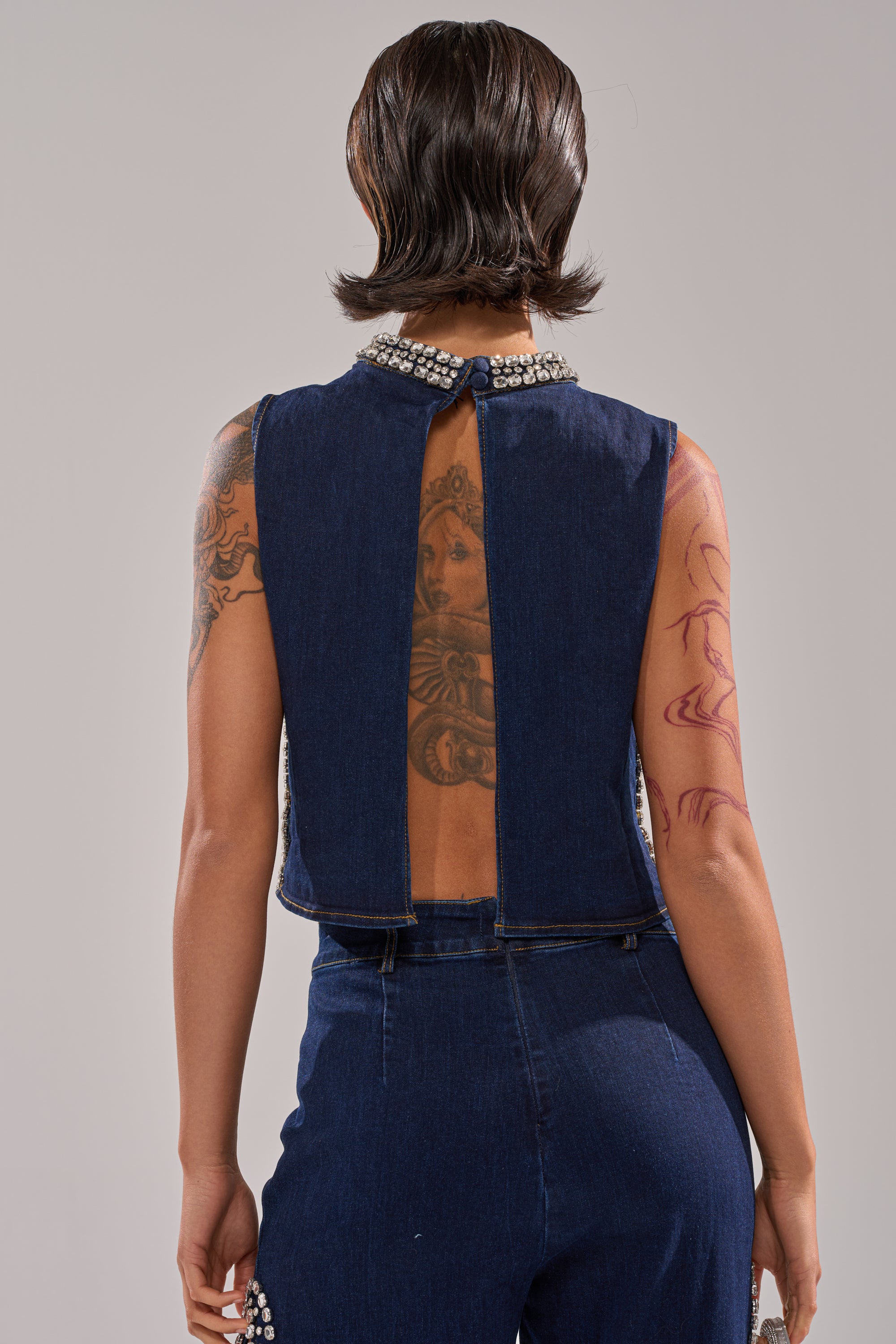A person with short dark hair and visible tattoos stands facing away, wearing the POPPIN OUT RHINESTONE DENIM BLOUSE IN DARK WASH DENIM, paired with matching high-waisted denim pants featuring metallic details. 4