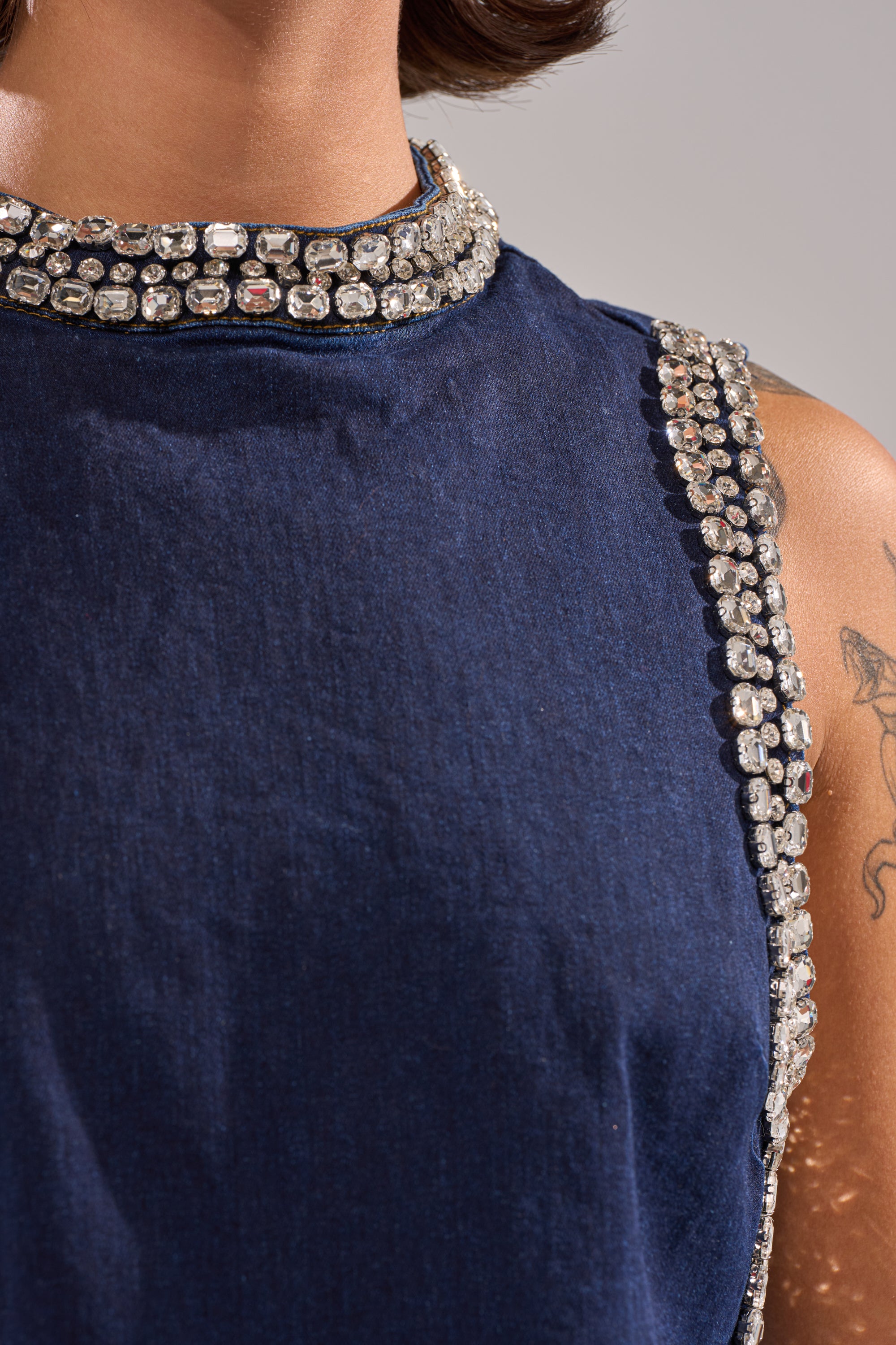 Close-up of a person wearing the POPPIN OUT RHINESTONE DENIM BLOUSE IN DARK WASH DENIM, featuring sparkling rhinestone embellishments on the collar and armholes. A tattoo is visible on their upper arm. 6