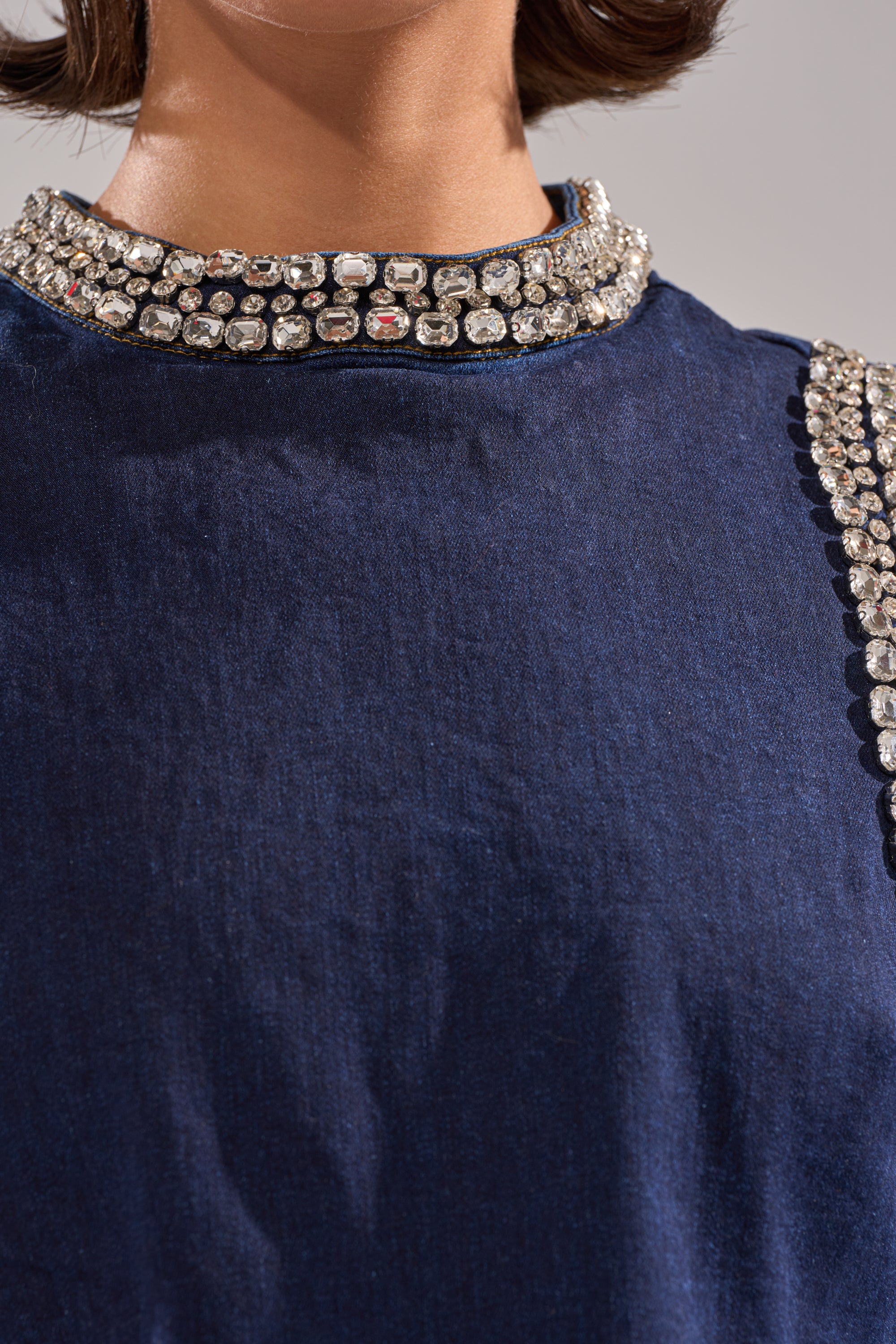 Close-up of a person in the POPPIN OUT RHINESTONE DENIM BLOUSE IN DARK WASH DENIM, featuring sparkling rhinestones along the collar and one shoulder. The smooth, elegant fabric is shown with the person's chin and part of their neck visible. 7