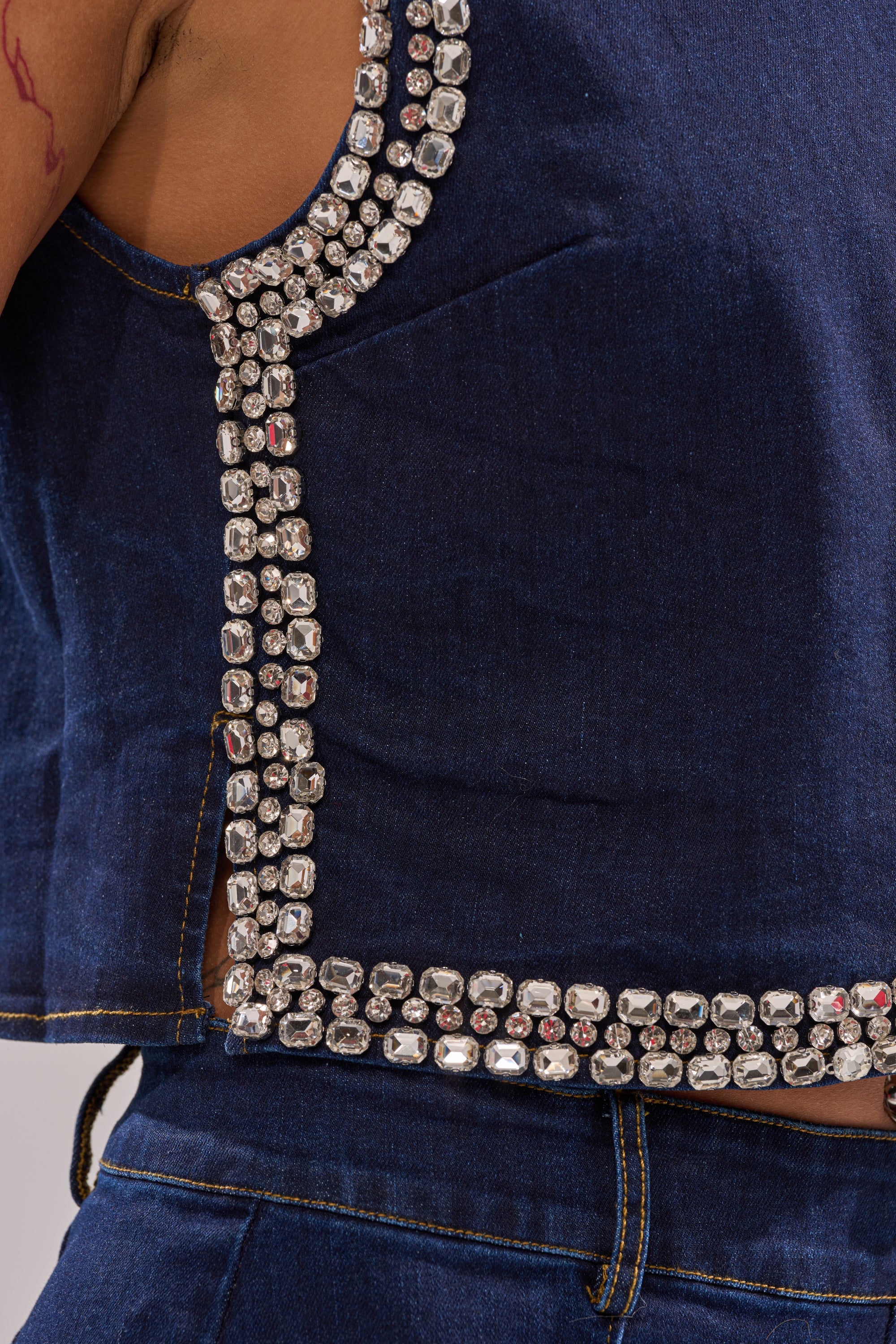 Close-up of a person wearing the POPPIN OUT RHINESTONE DENIM BLOUSE IN DARK WASH DENIM, featuring rows of large square silver rhinestones along the edges. A matching pant and a tattoo on their upper arm are also visible. 8
