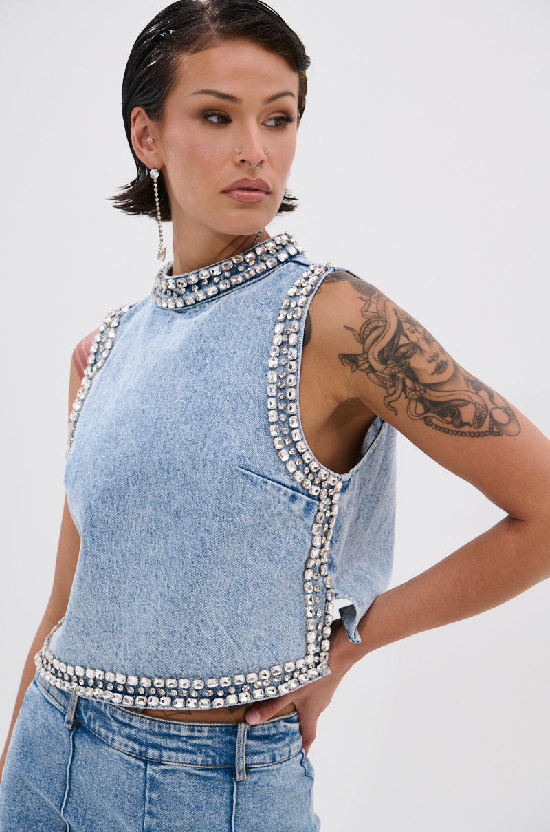 A woman with a tattoo on her upper arm and short, sleek hair wears the POPPING OUT RHINESTONE DENIM BLOUSE—sleeveless, high-neck, and light-wash—paired with matching denim bottoms and drop earrings.