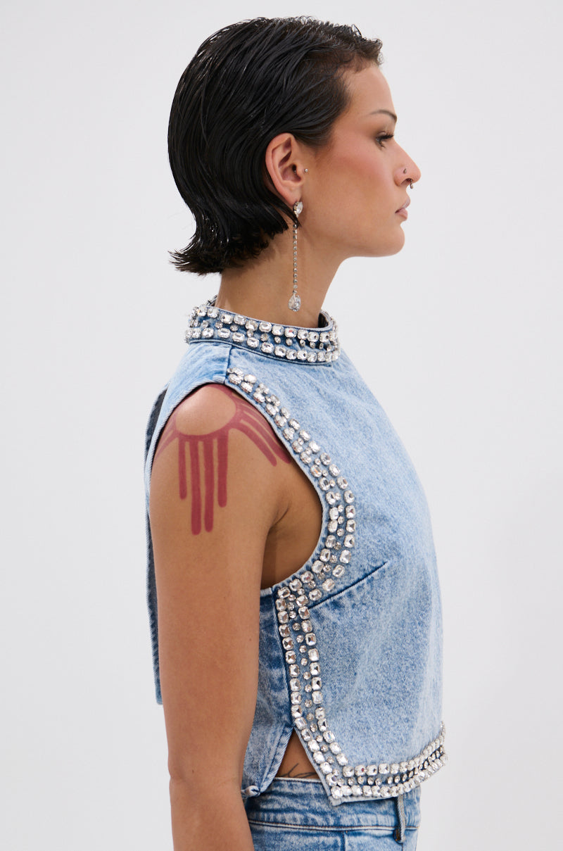 A person with short, slicked-back hair faces sideways, wearing the POPPING OUT RHINESTONE DENIM BLOUSE—a sleeveless, light blue, high-neck top with silver studs and a shoulder cutout revealing a red tattoo. They accessorize with a long earring.