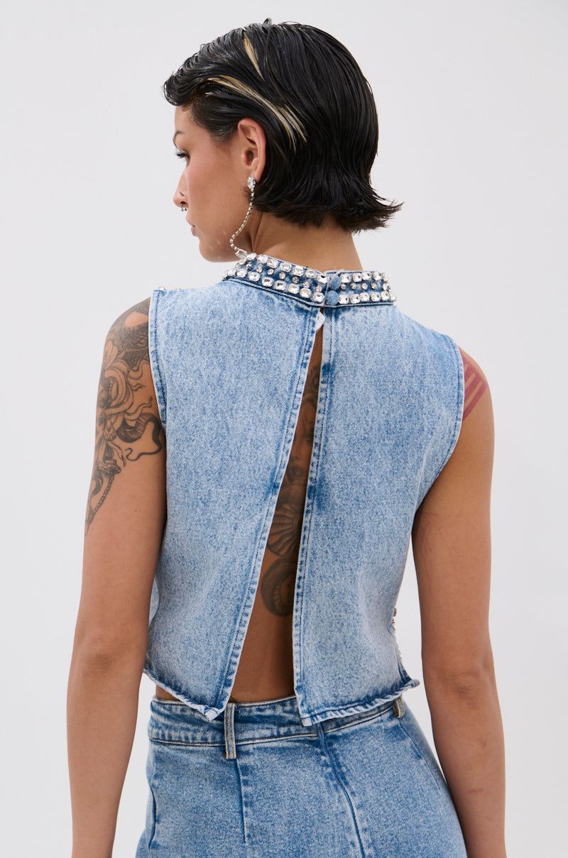 A person with short dark hair and tattoos is seen from behind, wearing the POPPING OUT RHINESTONE DENIM BLOUSE, a sleeveless high-neck denim top with pearl embellishments and an open back, paired with high-waisted jeans.