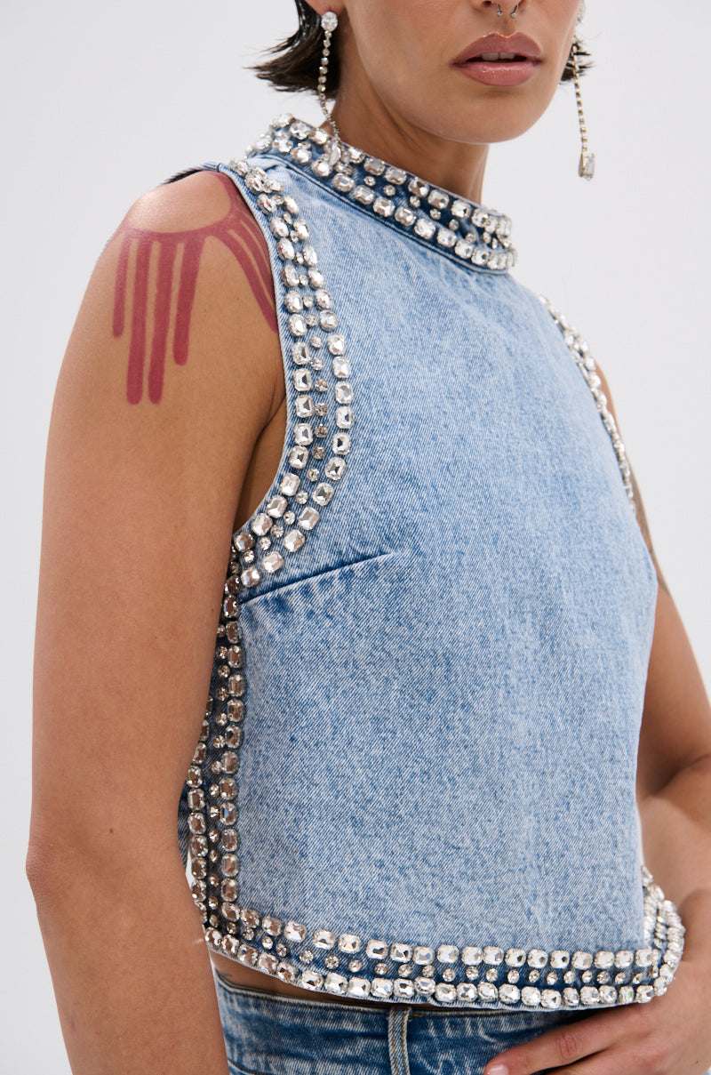 Wearing the POPPING OUT RHINESTONE DENIM BLOUSE—a sleeveless, high-neck light blue top with rhinestone embellishments—this person has a red, paint-drip tattoo on their shoulder and a long, dangling earring.