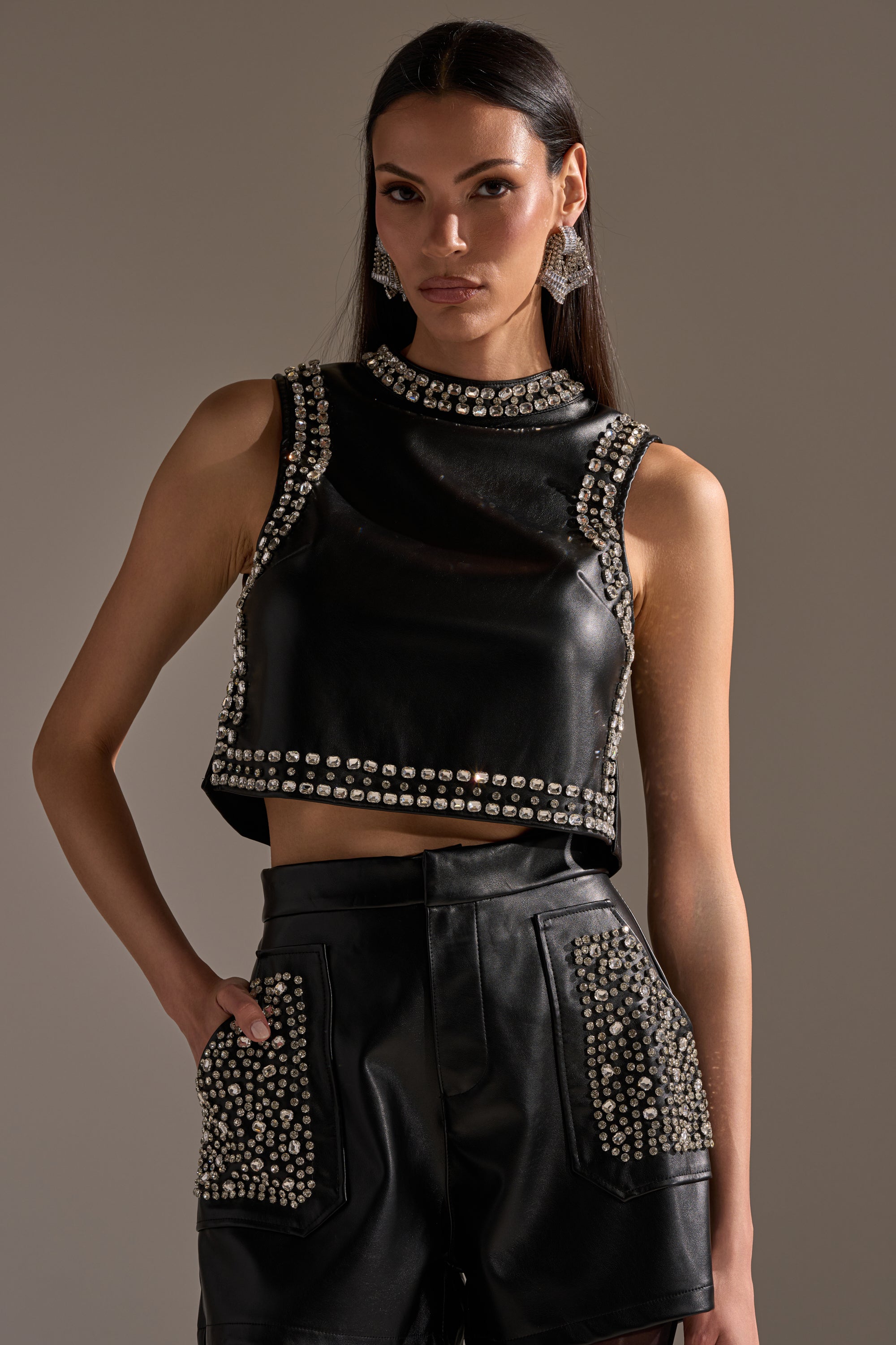 A woman poses against a plain background wearing the AKIRA Label DANCE THE NIGHT AWAY FAUX LEATHER RHINESTONE BLOUSE with high-waisted shorts, featuring silver studs, long straight hair, statement earrings, and one hand in her pocket. 0