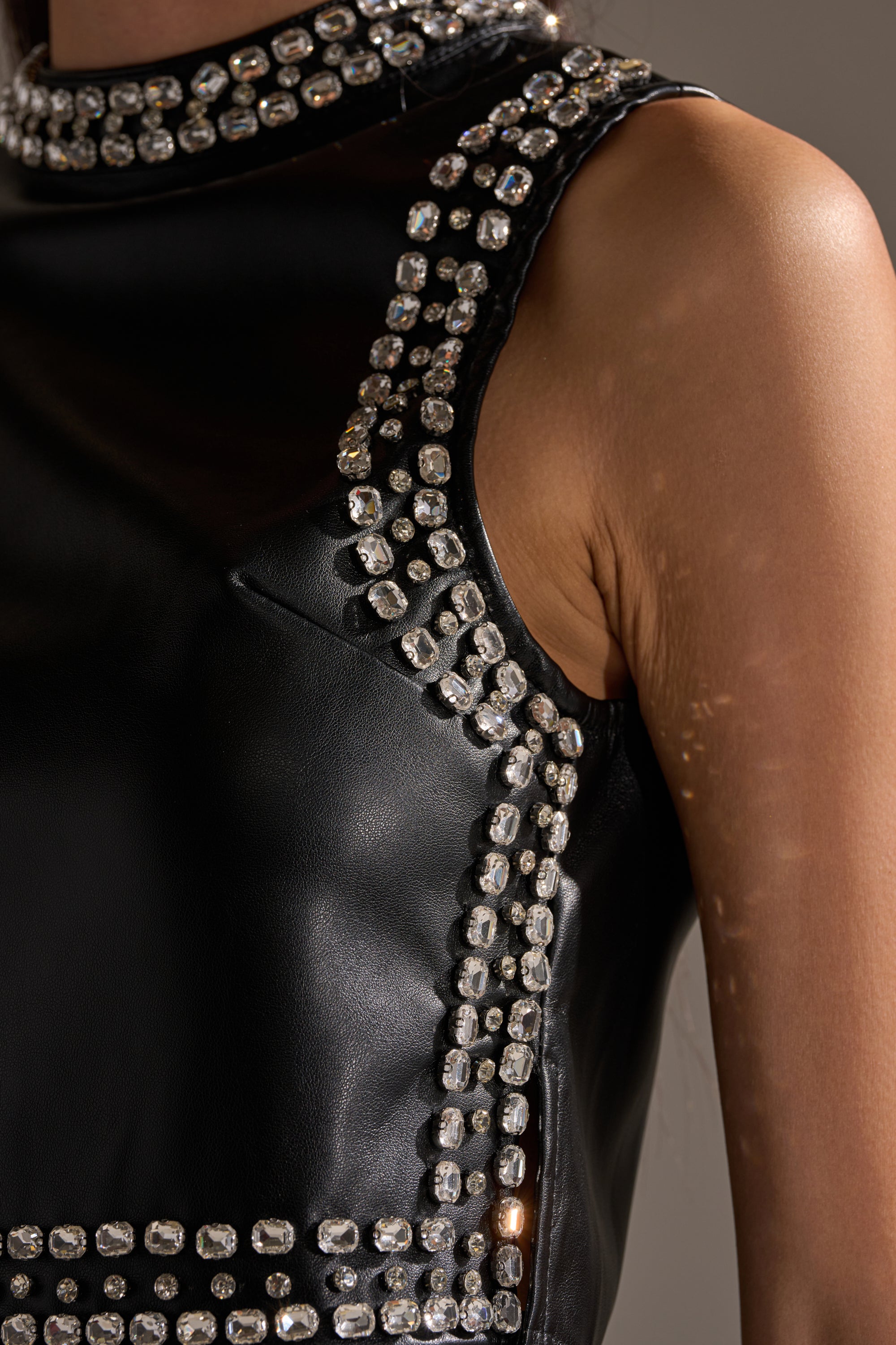 A person with medium-brown skin wears the DANCE THE NIGHT AWAY FAUX LEATHER RHINESTONE BLOUSE, a sleeveless mock neck top adorned with rows of square, clear rhinestones along the edges and seams. 9