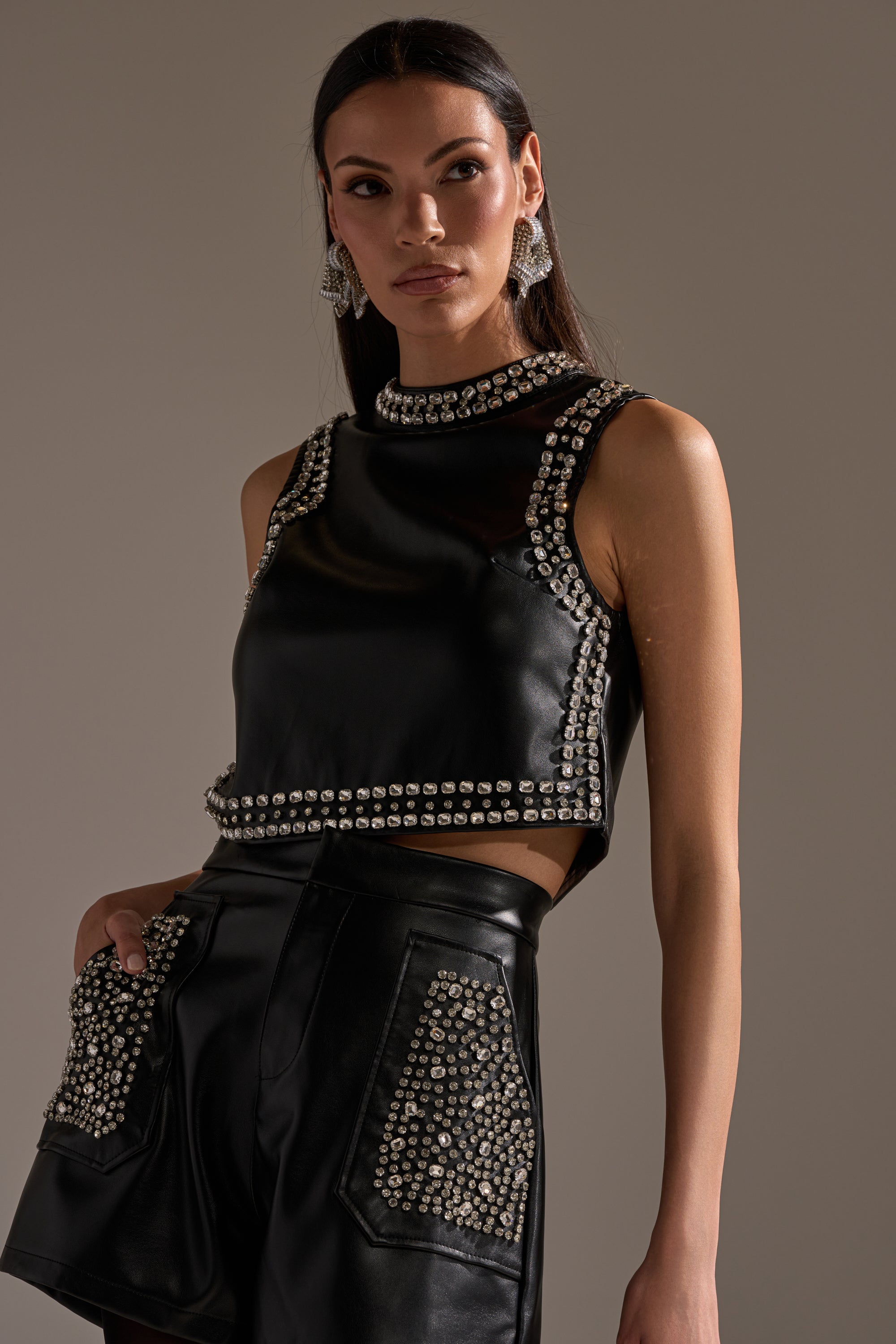 A woman stands confidently against a neutral background in the DANCE THE NIGHT AWAY FAUX LEATHER RHINESTONE BLOUSE, accessorized with statement earrings and long dark hair. 1