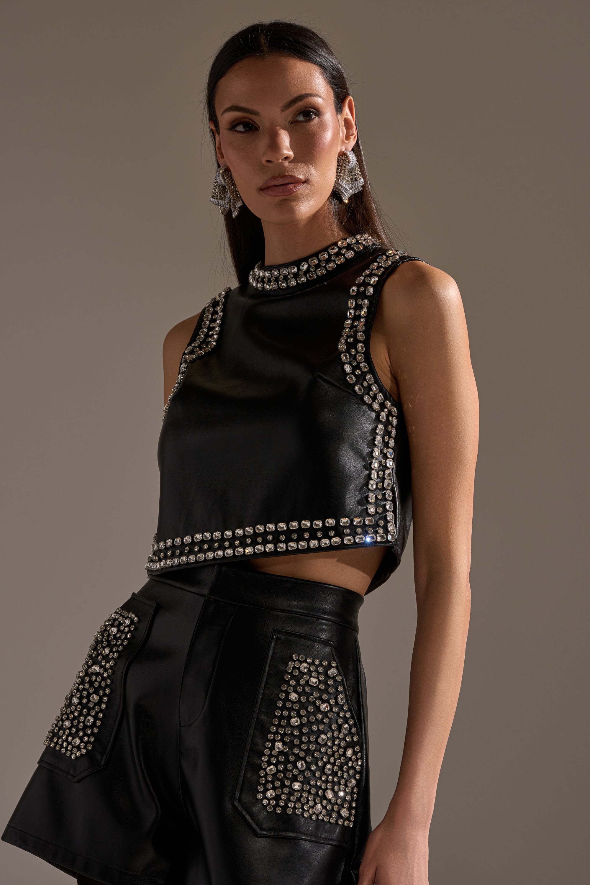 A woman with long dark hair and statement earrings poses confidently in the DANCE THE NIGHT AWAY FAUX LEATHER RHINESTONE BLOUSE—a sleeveless, mock neck black crop top decorated with silver studs—paired with matching high-waisted shorts. 2