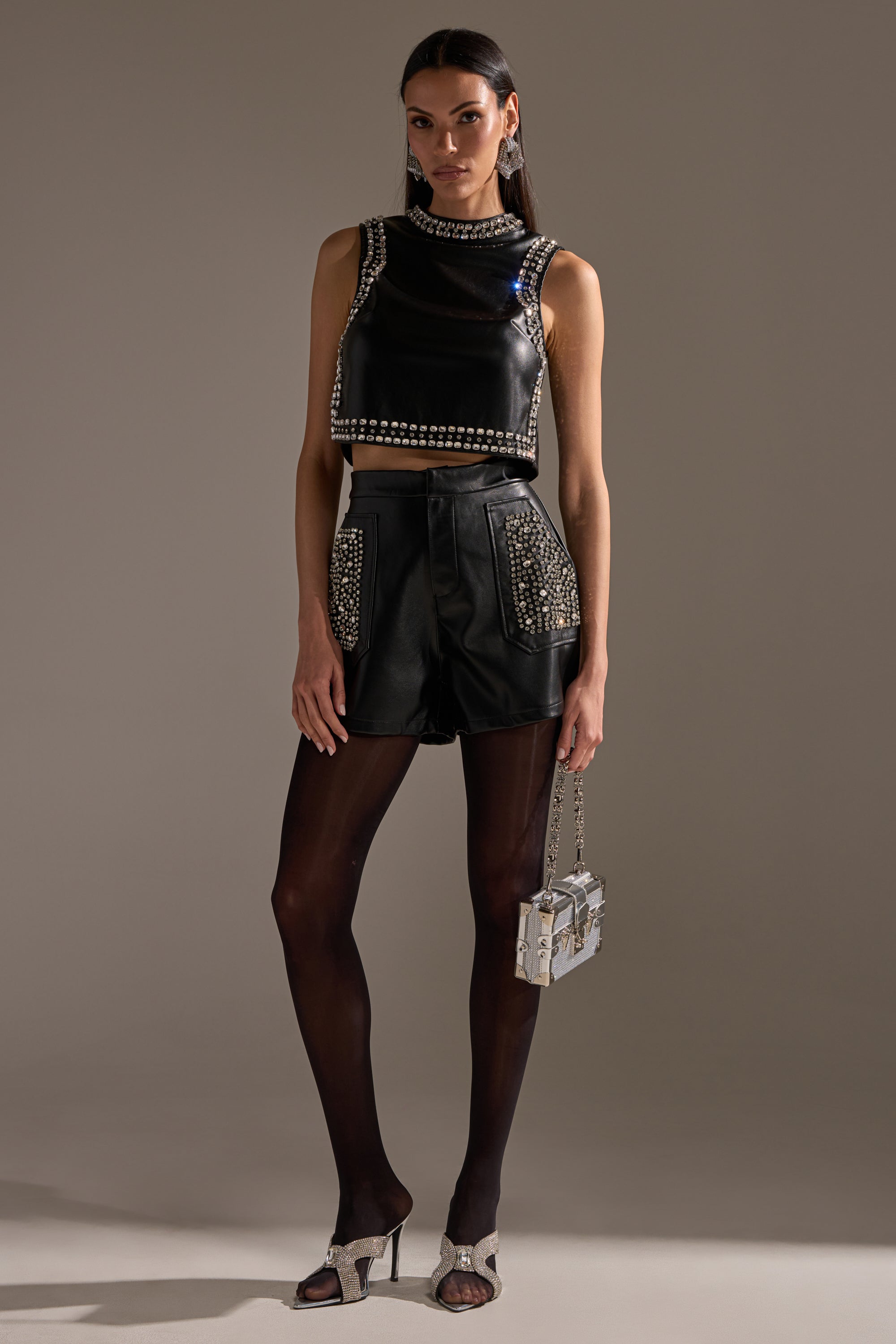 A woman poses against a plain background in the DANCE THE NIGHT AWAY FAUX LEATHER RHINESTONE BLOUSE, styled with matching high-waisted shorts, sheer black tights, open-toe heels, and a small metallic handbag. 5