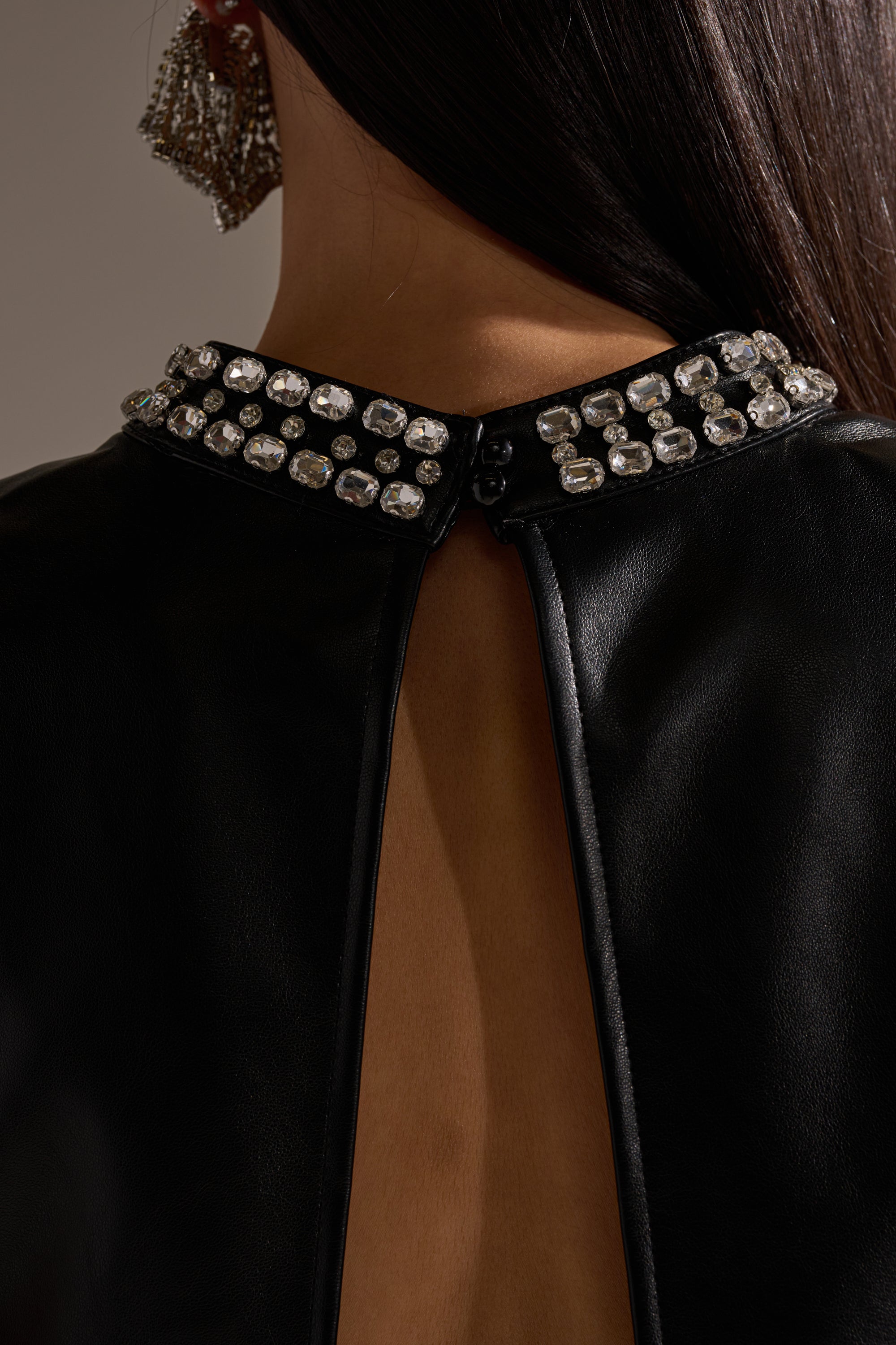 Close-up of a person in the DANCE THE NIGHT AWAY FAUX LEATHER RHINESTONE BLOUSE, featuring a sleeveless mock neck, open back, and collar with large sparkling rhinestones. The person has long dark hair and wears a dangling beaded earring. 8