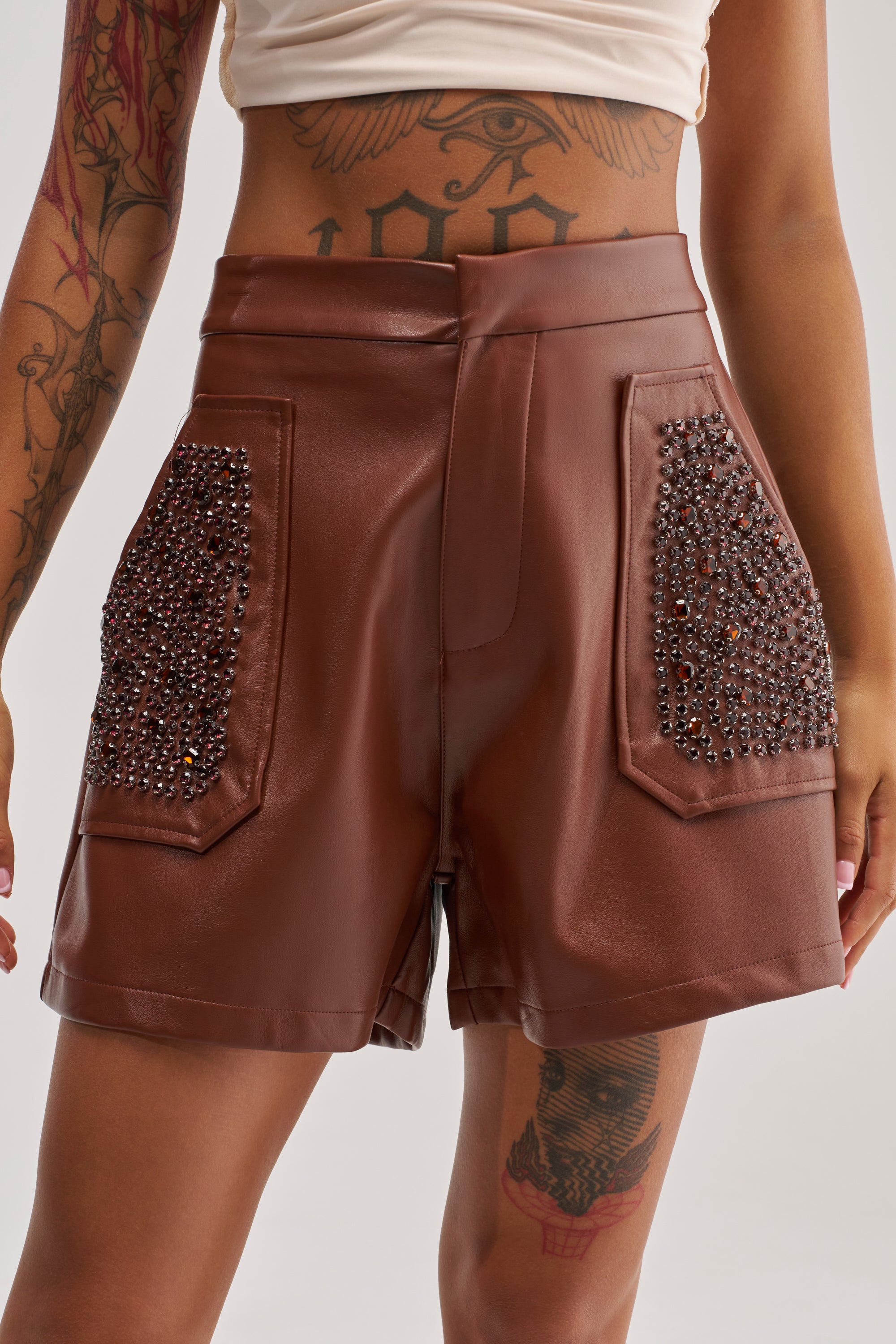 Wearing the HOW ABOUT THIS FAUX LEATHER RHINESTONE SHORT—high-rise, brown faux leather shorts featuring large rhinestone-adorned front pockets—a person pairs them with a cream crop top, displaying tattoos on their arm and thigh.