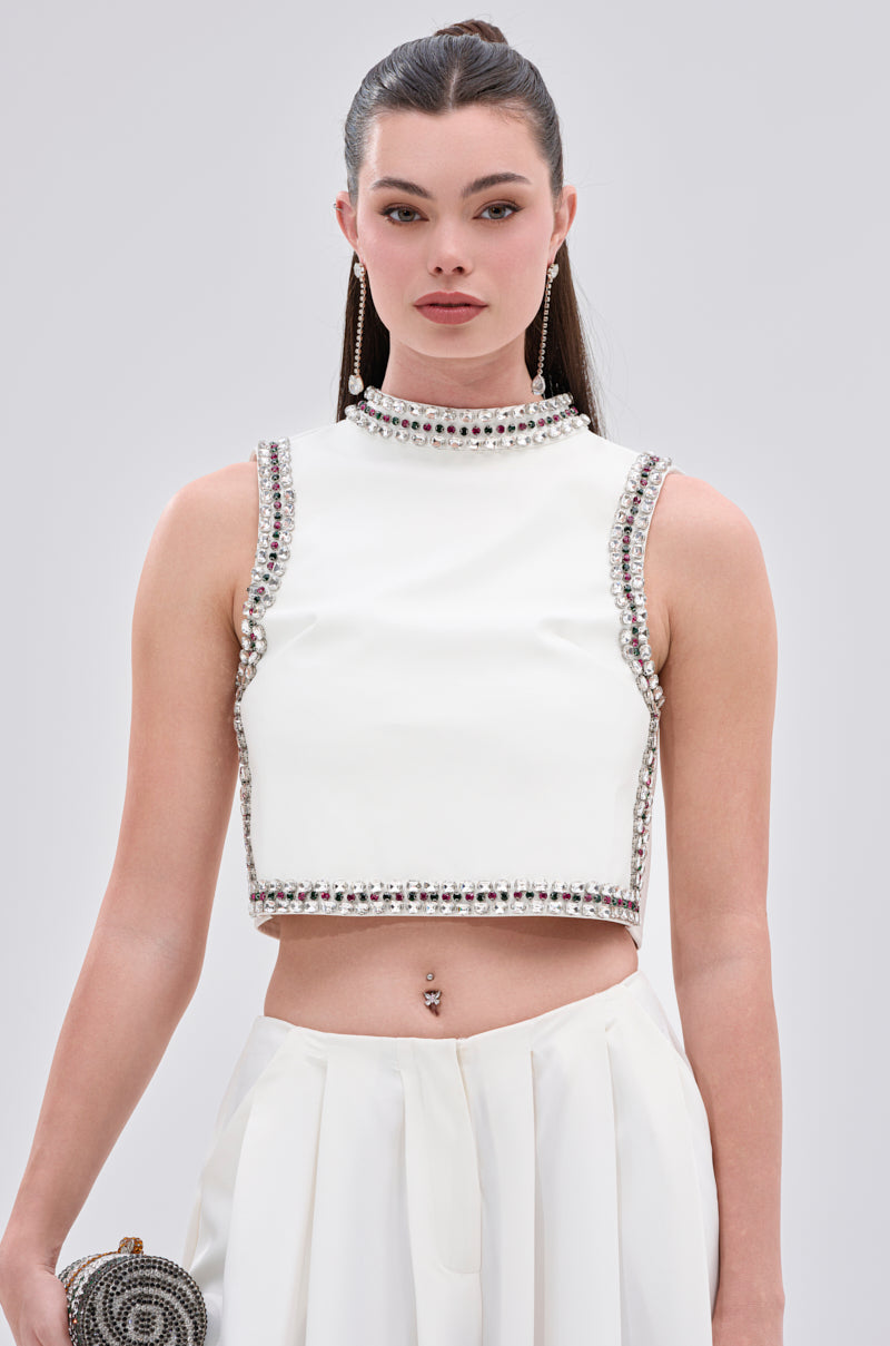 A woman in the SPARKLE QUEEN BLOUSE with rhinestone embellishments and matching high-waisted pants holds a small clutch against a plain light gray background.