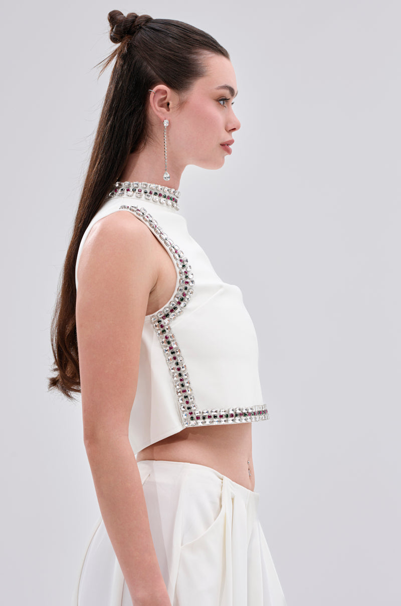 A woman with long, dark hair styled half-up models the SPARKLE QUEEN BLOUSE—a sleeveless white crop top and skirt adorned with rhinestones—posing in profile against a plain light background.