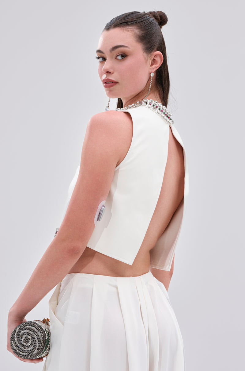 A woman in the SPARKLE QUEEN BLOUSE, featuring an open-back design and rhinestone-embellished collar, holds a beaded clutch and wears long earrings with a sleek updo, gazing over her shoulder against a plain backdrop.