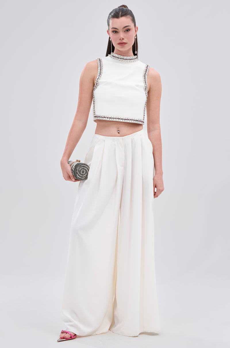 A woman wears the SPARKLE QUEEN BLOUSE, a sleeveless white faux leather top with rhinestone embellishments, paired with wide-leg white pants, a silver clutch, and pink sandals against a plain light background.
