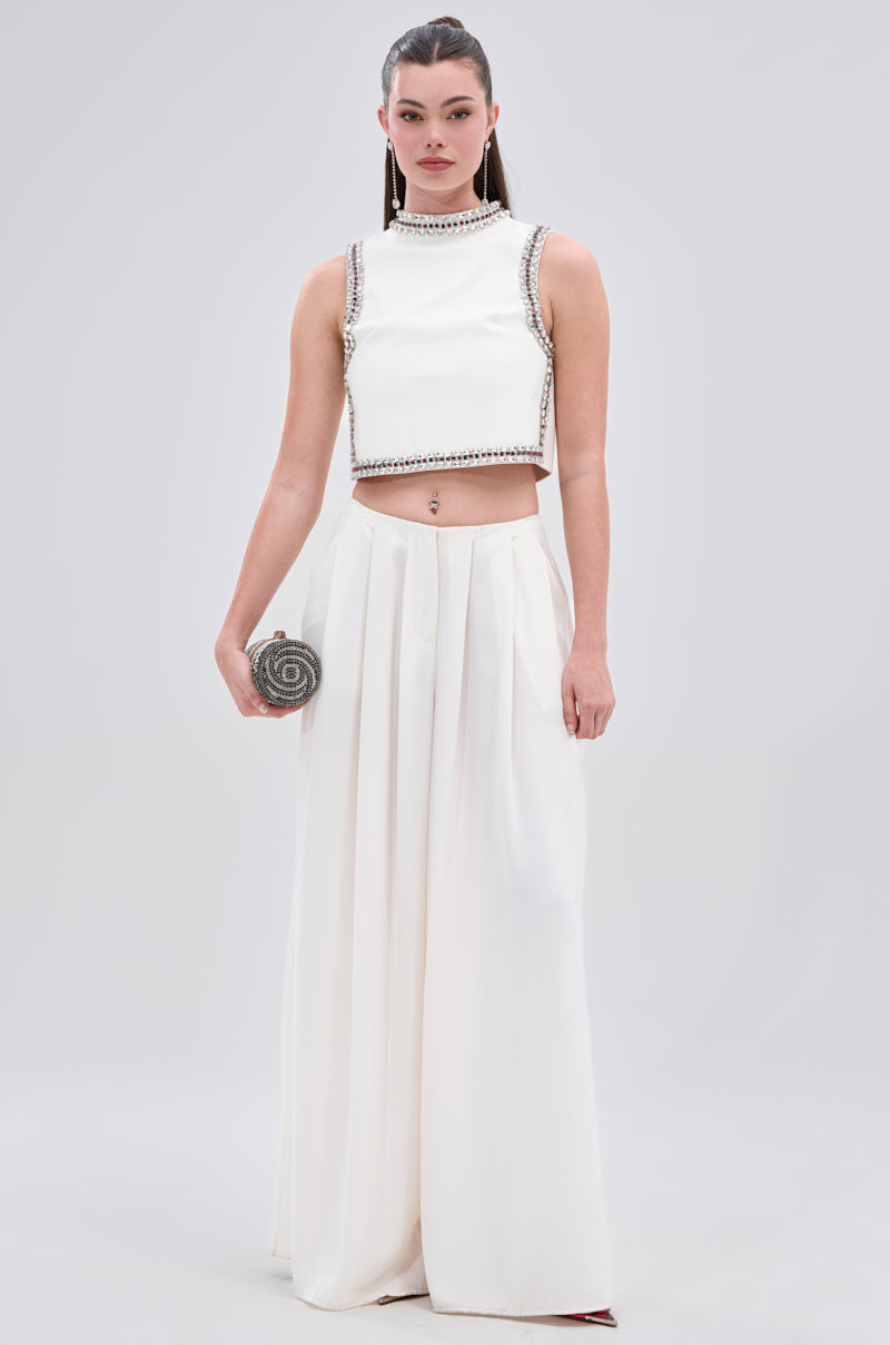 A woman faces forward wearing the SPARKLE QUEEN BLOUSE, a sleeveless white crop top with rhinestone embellishments, paired with a high-waisted white skirt and holding a silver clutch against a light gray background.