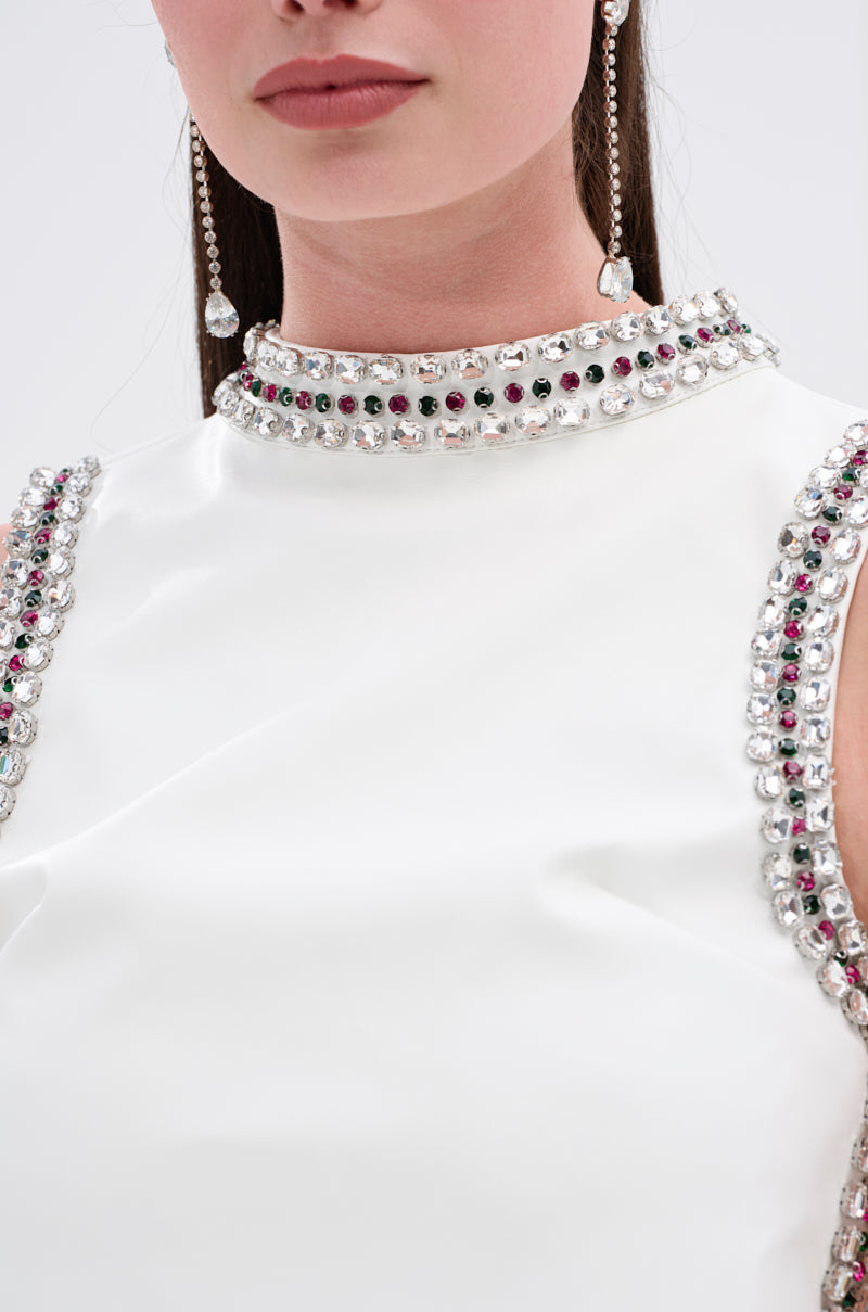 A close-up shows a woman wearing the SPARKLE QUEEN BLOUSE, a white sleeveless top with rows of clear and dark gemstones on the collar and armholes, paired with long gemstone earrings. Only her lower face and upper torso are visible.