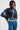 A woman stands against a plain white background wearing the GALAXY BLOUSE—an avant-garde, pleated high-neck top with a blue-and-black galaxy print—paired with high-waisted blue jeans, a chunky silver chain belt, and silver earrings.