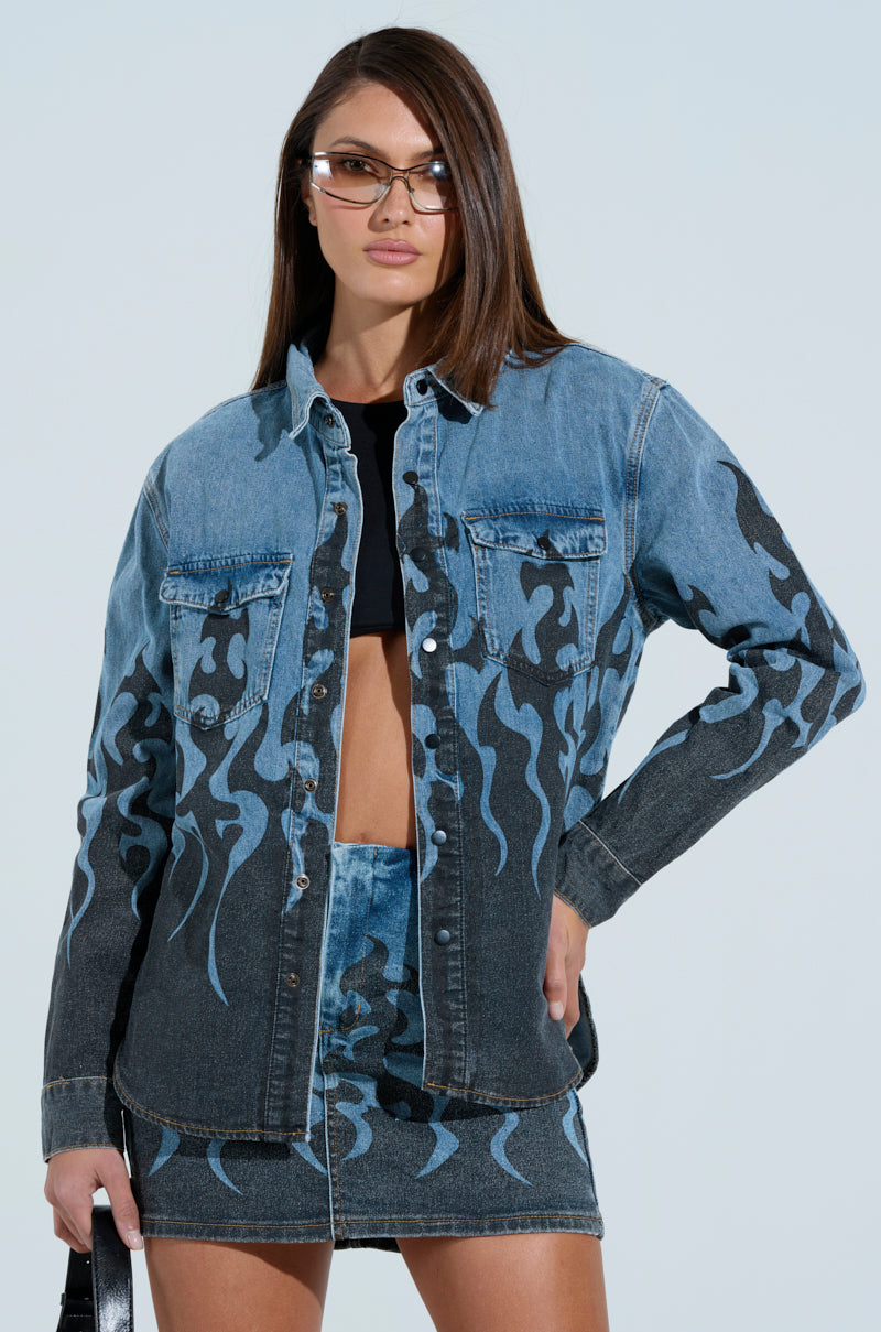 A person showcases an oversized silhouette with the FIRE TO MY FLAME DENIM SHIRT featuring black flame detailing, paired with a chic denim skirt. Long dark hair and sunglasses add flair, while a sleek black top peeks from beneath, all set against a serene light blue background. 0