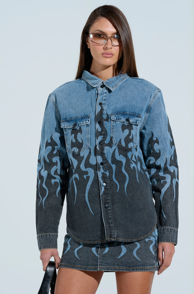 A woman with glasses models the FIRE TO MY FLAME DENIM SHIRT, which features snap buttons and vivid blue flame detailing, paired with a matching denim skirt. She carries a black handbag against a light blue backdrop. 2