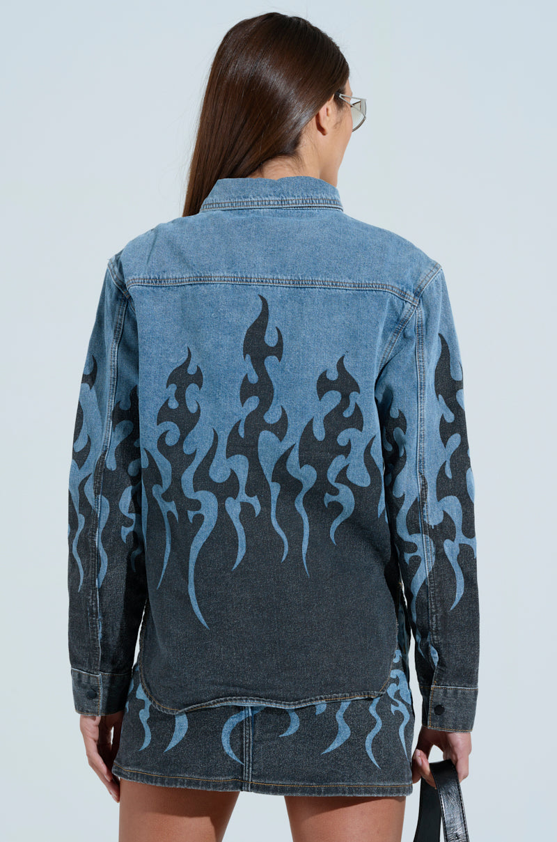 A person with long hair wears sunglasses and the FIRE TO MY FLAME DENIM SHIRT, complete with blue flame detailing on the back and sleeves. They pair it with a matching snap-button denim skirt and hold a black bag against a muted, light background. 4