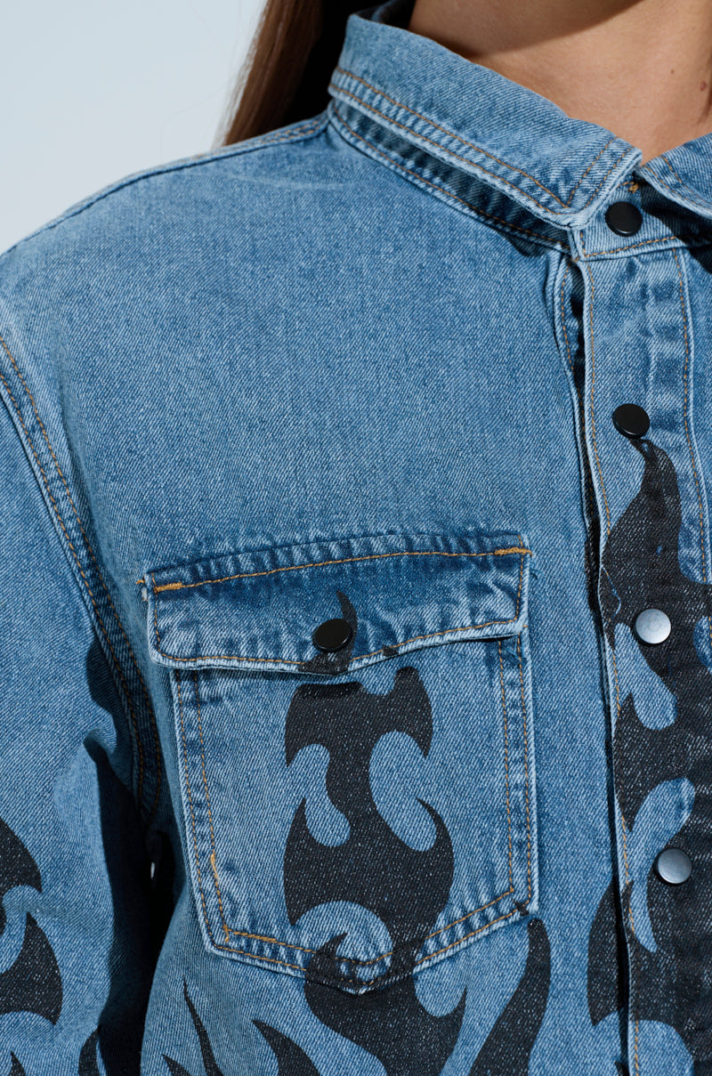Close-up of the FIRE TO MY FLAME DENIM SHIRT, an oversized denim jacket with black flame detailing. It has a front chest pocket with a buttoned flap and sleek black buttons down the front, all set against a light neutral background. 7