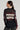 A person with long, dark hair tied in a low ponytail is seen from behind. They are wearing an AMERICANA GIRL LONG SLEEVE SHIRT made of black sheer fabric featuring red and beige stripes and the text "MERION" printed on the back. Completing their look are large, round hoop earrings.